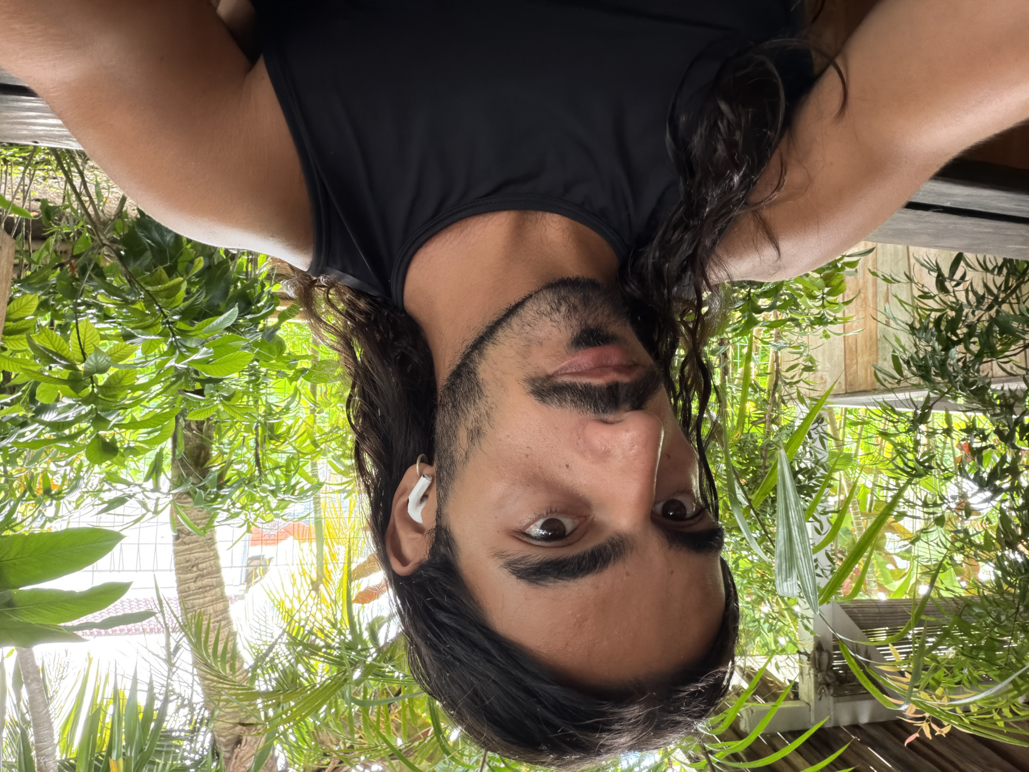 A man with long dark hair and a beard is pictured in an upside-down selfie. He is wearing a black sleeveless shirt and an earpiece. The background features tropical foliage, including palm trees and lush greenery. The location is determined to be North Kuta, Indonesia.