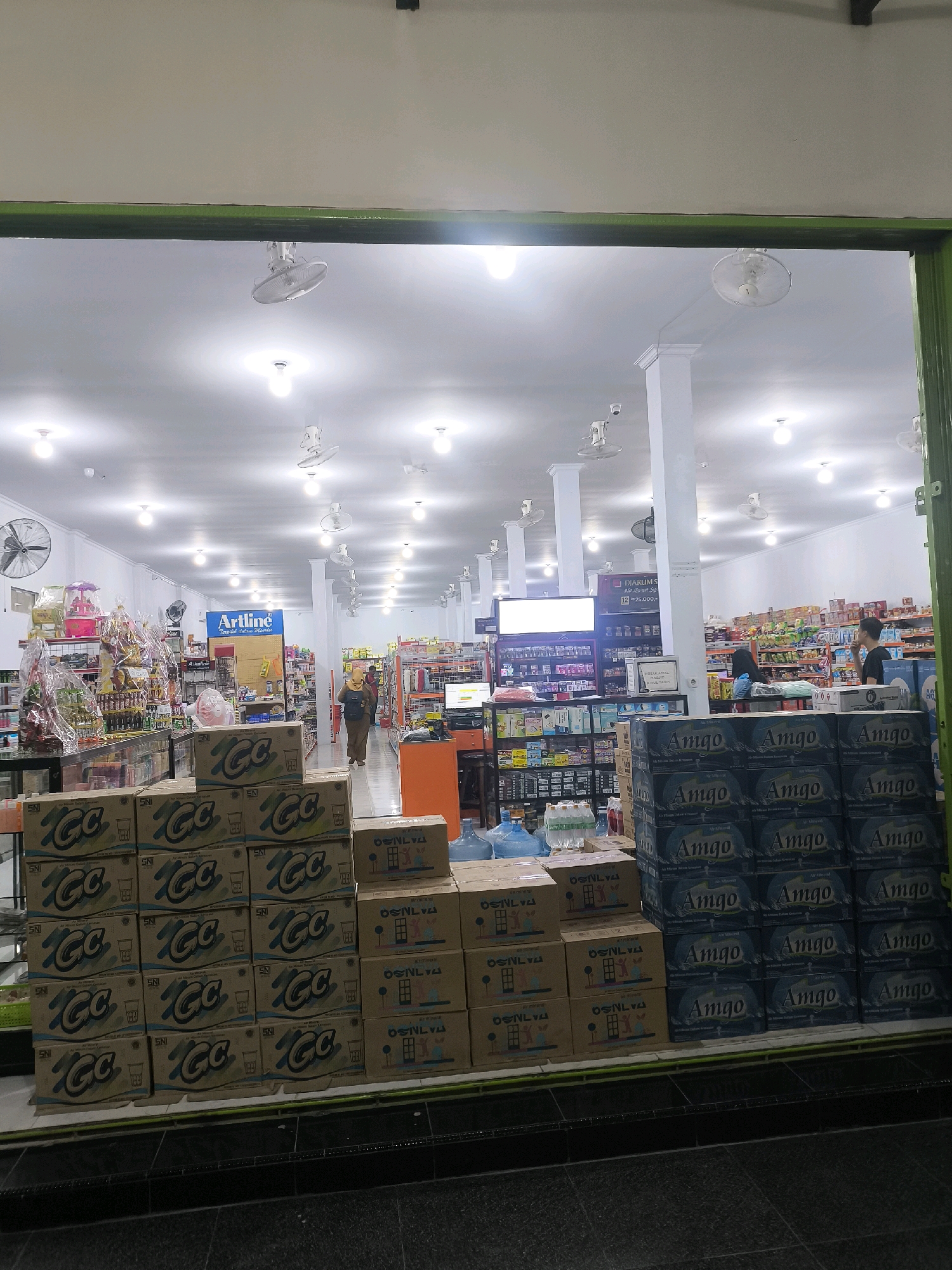 This image depicts the interior of a grocery store or convenience store, likely located in Pilolahuwa, Indonesia. The store is well-lit with ceiling lights and illuminated by natural light filtering in from the entrance. Stacks of boxed beverages are prominently displayed in the foreground, with "CC" branding visible on some and "Amigo" branding on others. To the right, there are large blue water jugs. The shelves in the background are stocked with various products, including packaged goods and household items. Several ceiling fans are visible, suggesting a warm climate. A few people are present in the store, including a woman in a headscarf walking down an aisle and a person standing near a checkout counter. The overall impression is that of a typical retail environment with abundant merchandise.
