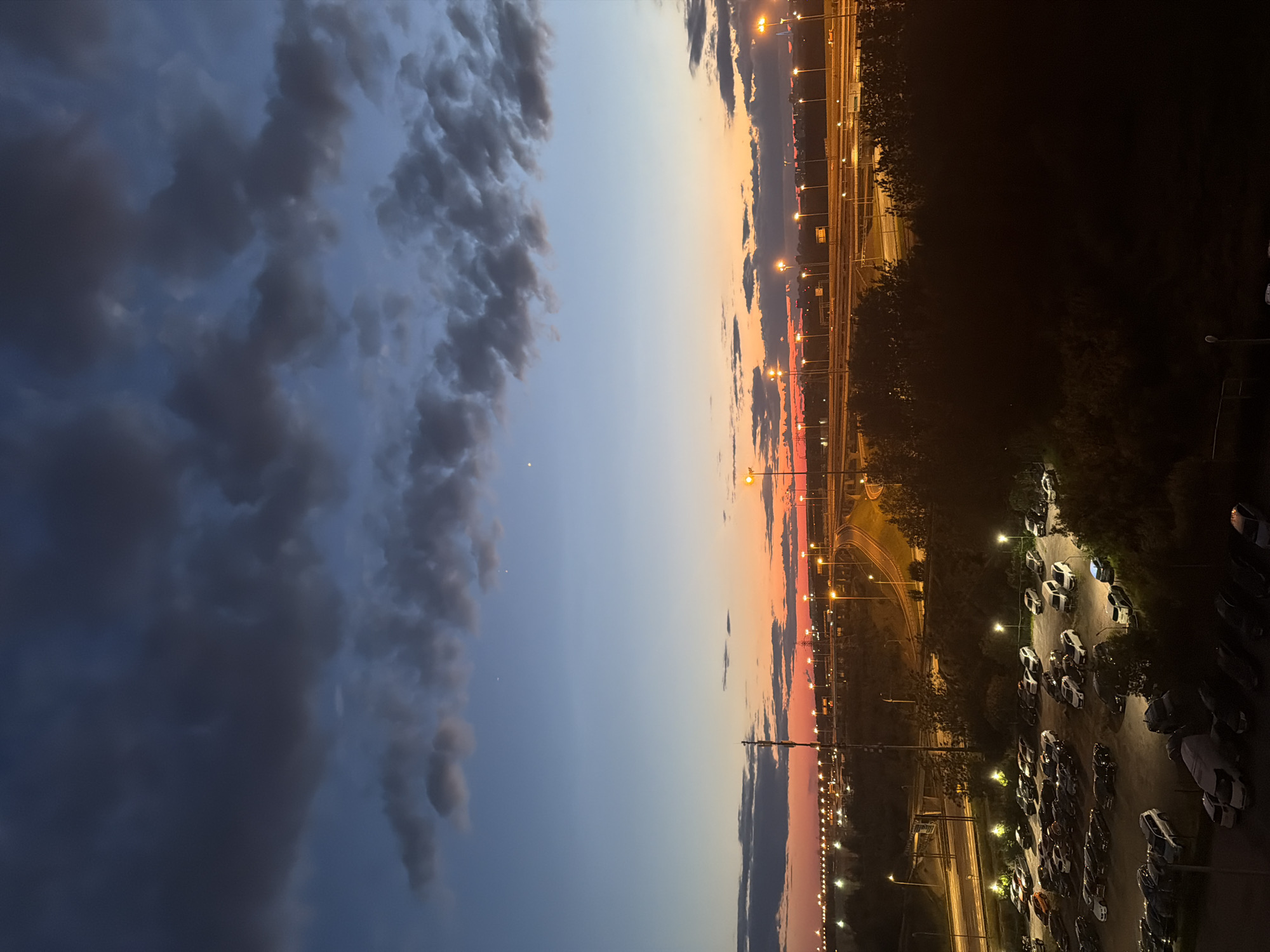 The image captures a twilight scene over a city, with a dramatic sky transitioning from deep blues and grays at the top to fiery oranges and reds near the horizon. Dark, textured clouds are scattered across the upper portion of the sky, while a faint glow from the setting sun is visible just above the distant city lights and highway infrastructure.
The foreground is dominated by a large, dimly lit parking lot filled with numerous cars of various makes and models, suggesting a commercial or industrial area. The vehicles are parked in neat rows, and their headlights or interior lights are not illuminated. Some areas of the parking lot are shrouded in shadow. Adjacent to the parking lot, dense foliage and trees are visible, obscuring further details of the ground and some of the parking lot's perimeter.
In the mid-ground, a complex network of highways and overpasses is illuminated by rows of streetlights, casting a warm, yellow glow on the roadways and surrounding landscape. The highway appears to be a multi-lane thoroughfare, with traffic potentially present but not clearly discernible due to the low light conditions. A tall telecommunications tower stands prominently on the left side of the frame, silhouetted against the twilight sky.
The background reveals the sprawling urban landscape with a multitude of lights, indicating a developed area. The overall impression is that of a city on the cusp of night, with the artificial lights beginning to dominate the natural light of the fading day. The scene is devoid of any visible human figures.