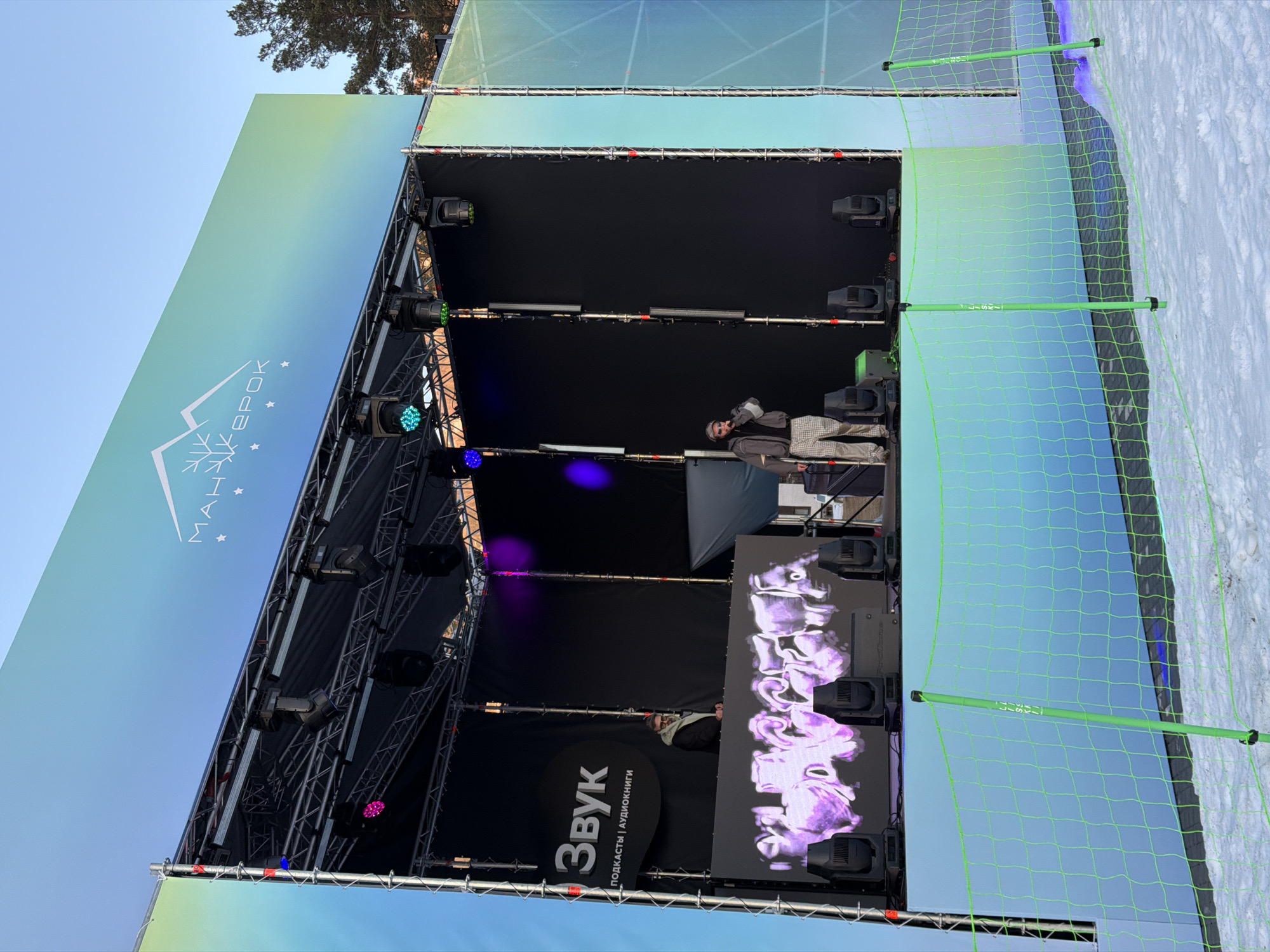 The image shows an outdoor stage setup in Озёрное, Russia. The stage is equipped with lighting fixtures, a large LED screen displaying graphics, and sound system elements. A person is standing on the stage, possibly adjusting equipment. The stage is surrounded by a net fence, and snow is visible on the ground, suggesting a winter event. The backdrop includes signage in Russian.