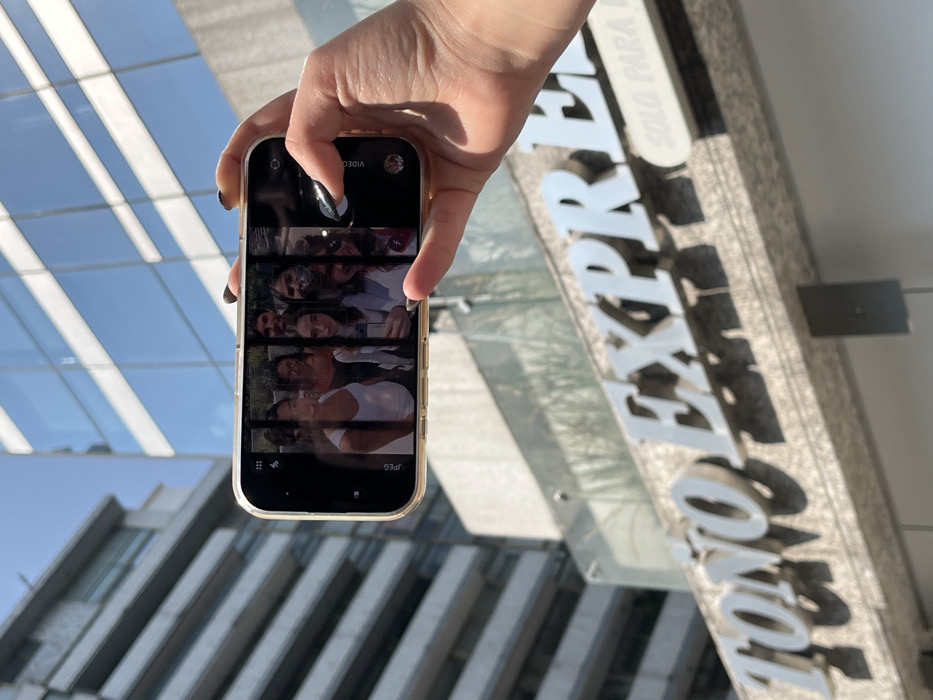A hand holds a cell phone displaying a collage of faces. The phone is framed against the backdrop of a modern building with glass windows. To the right, a sign with the words "TONOS EXPRESS" in large silver letters is partially visible. The overall composition creates an interplay between personal technology, architecture, and branding.