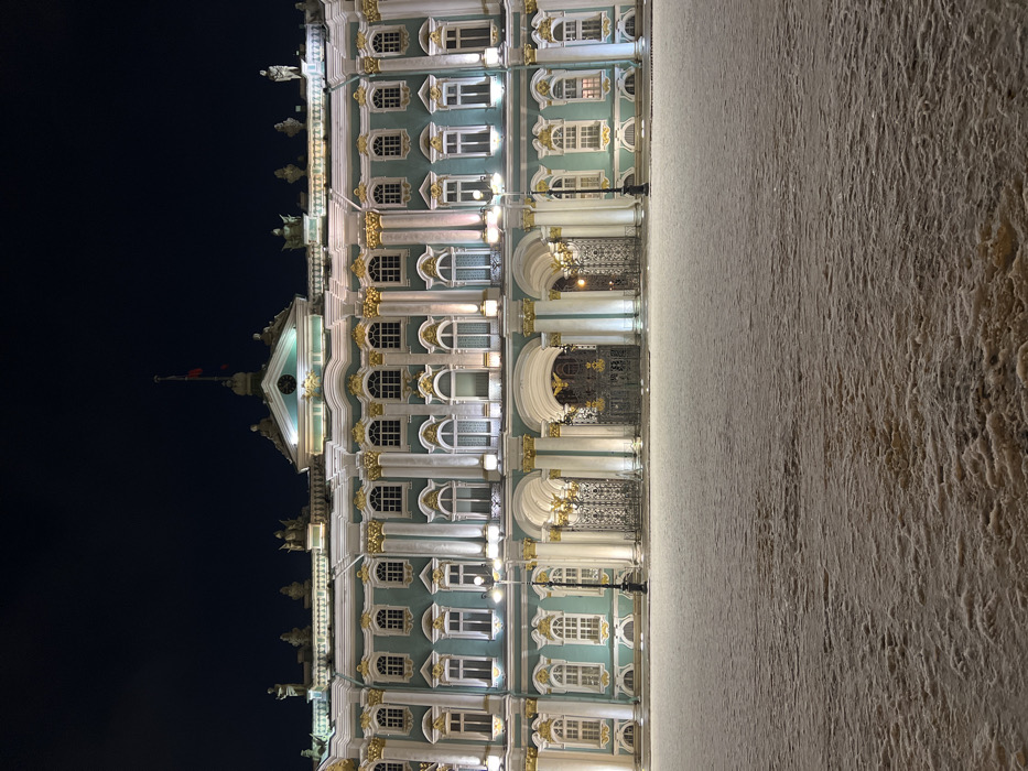 An ornate, multi-storied building is dramatically illuminated at night. The building's facade features rows of windows adorned with gilded accents. A flag waves atop the building's peak. To the left of the building is a dark sky, contrasting with the bright facade. Adjacent to the building on the right is a stark, textured wall or pavement area, lit dimly. The image is oriented vertically.