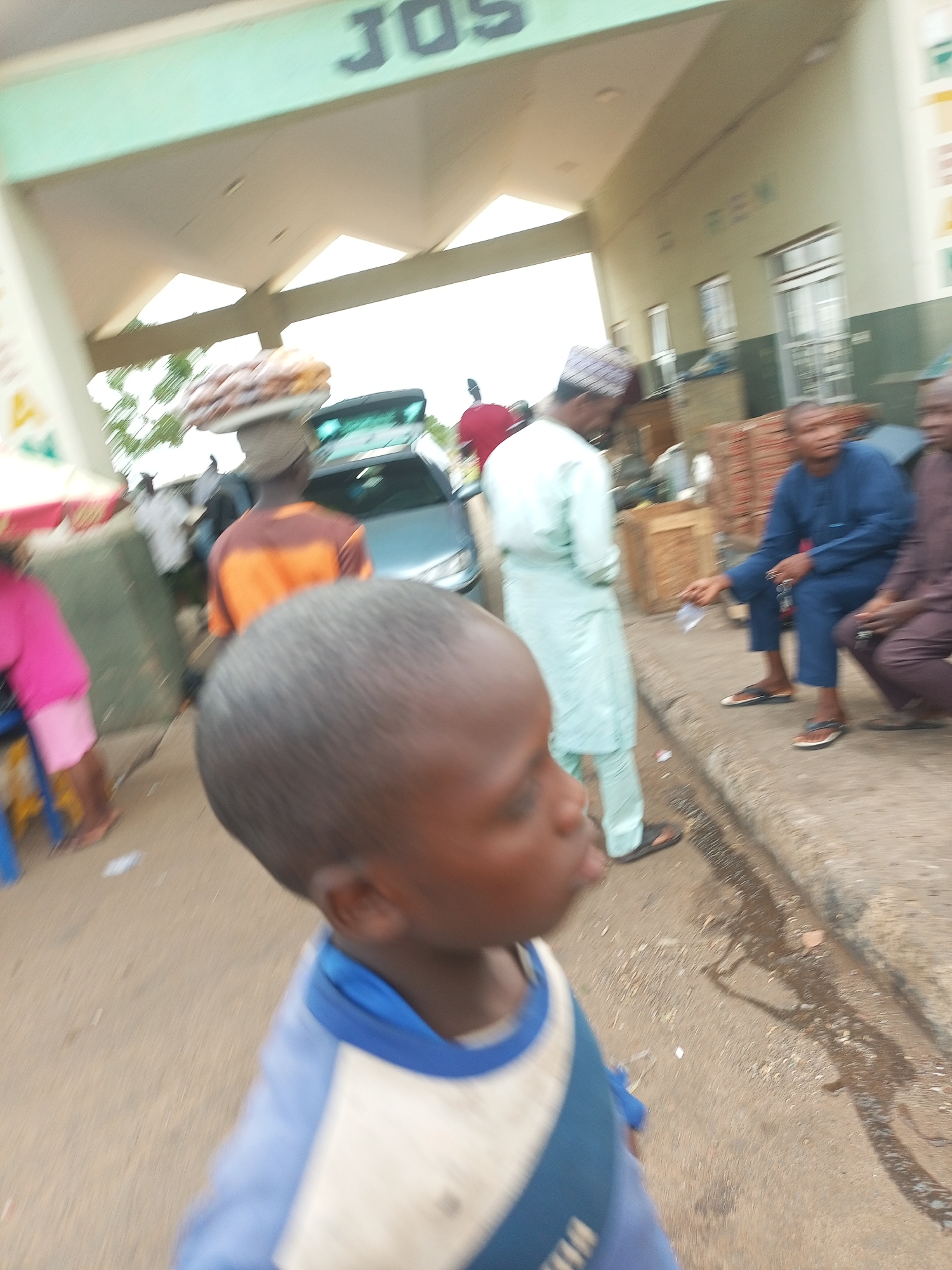The image depicts a street scene in Gombe, Nigeria, likely near a transportation hub or market given the presence of vehicles and people carrying goods. A young boy in the foreground, facing away from the camera, wears a blue and white shirt. Behind him, a man in an orange and brown striped shirt carries a large bundle on his head, with a car and other individuals in the background. To the right, a man in a blue robe is seated on what appears to be a bench or curb, interacting with another person. The environment is an outdoor urban setting with a paved surface and a building with signage that reads "JOS REM." The lighting suggests it is daytime.