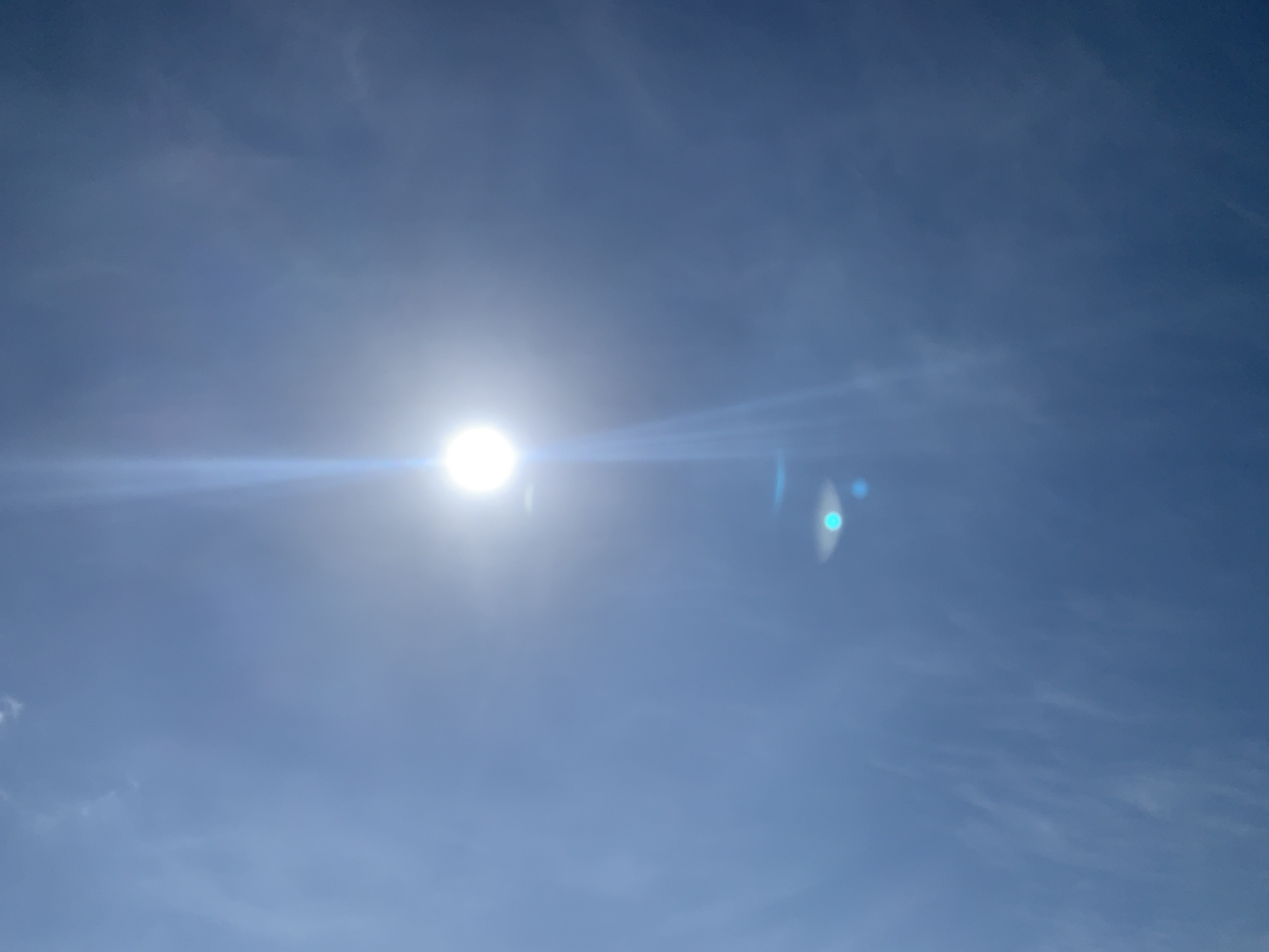 The image shows a bright sun in a clear blue sky with some wispy clouds. There are noticeable lens flares, including streaks of light and circular or oval reflections, one of which has a bluish tint. The scene is likely outdoors and depicts daylight conditions. There are no people, subjects, objects, actions, or interactions visible in the image that would suggest a specific location within Kuru Karama, Nigeria, or any other city. The image is purely a depiction of the sky and the sun.