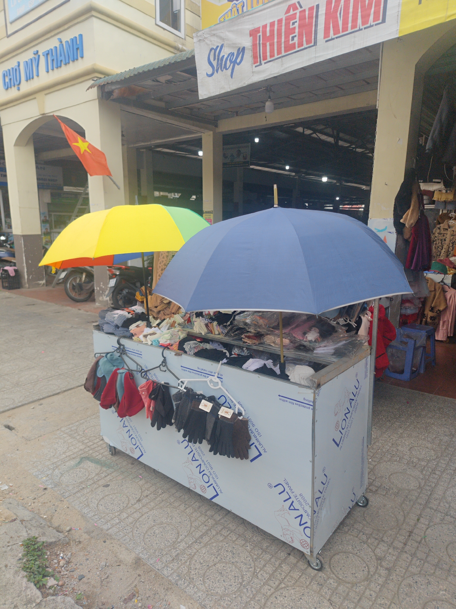 This photograph depicts a small, mobile retail stall in Cam Ly - Đà Lạt, Vietnam. The stall, constructed from aluminum composite panels branded "LIONALU," is set up on a paved pedestrian area. It showcases various clothing items, including gloves and other garments, displayed under two umbrellas: one is a bright yellow and green, while the other is a solid blue. These umbrellas serve to protect the merchandise from the elements.
Behind the stall, a building with signage reading "CHỢ MỸ THÀNH" (My Thanh Market) can be seen. The Vietnamese flag is also prominently displayed near the market entrance. Further in the background, a motorbike is parked. The scene suggests a typical small business operating in a local market setting.