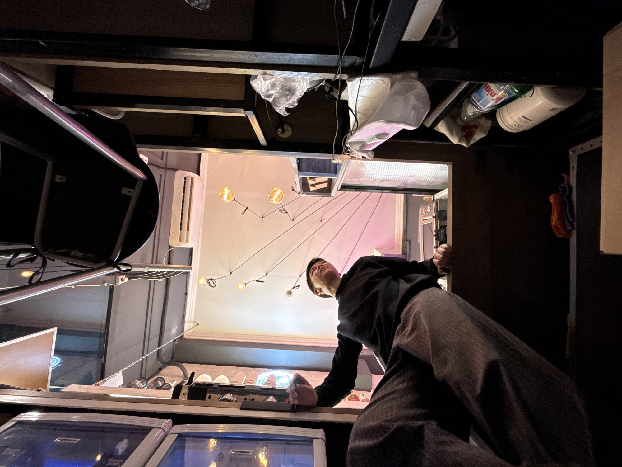 A young Asian man wearing a black long-sleeved shirt and brown pinstriped pants sits on a wooden surface, his legs dangling down. He is looking upwards and to his right. In the background, a series of lights are strung along wires, creating a zig-zag pattern. To the left of the man, a glass display case shows some objects inside. To the right, shelves hold various items, including plastic bags and cleaning supplies. An air conditioner is mounted high on the wall in the center background. The overall impression is that of an indoor space with a somewhat cluttered but functional aesthetic, possibly a shop or workshop. The image provides no specific clues to its location in Moscow, Russia.