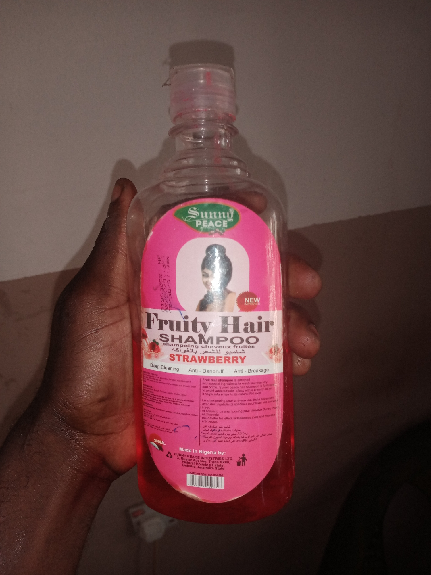An individual's hand is partially visible on the left side of the frame, holding a clear plastic bottle of "Sunny Peace Fruity Hair Shampoo" with a pink label. The label features an image of a woman with dark hair, the brand name "Sunny Peace" in green text, and "Fruity Hair SHAMPOO" in large, dark lettering. Below this, there are text descriptions in English, French, and Arabic, with "STRAWBERRY" prominently displayed. The text "NEW IMPROVED" is also visible on a pink sticker. The bottle contains a pink liquid. The background appears to be a plain, neutral-colored wall, suggesting an indoor setting. The lighting is dim, with a shadow cast by the bottle on the wall. The image appears to have been taken in Jalingo, Nigeria.