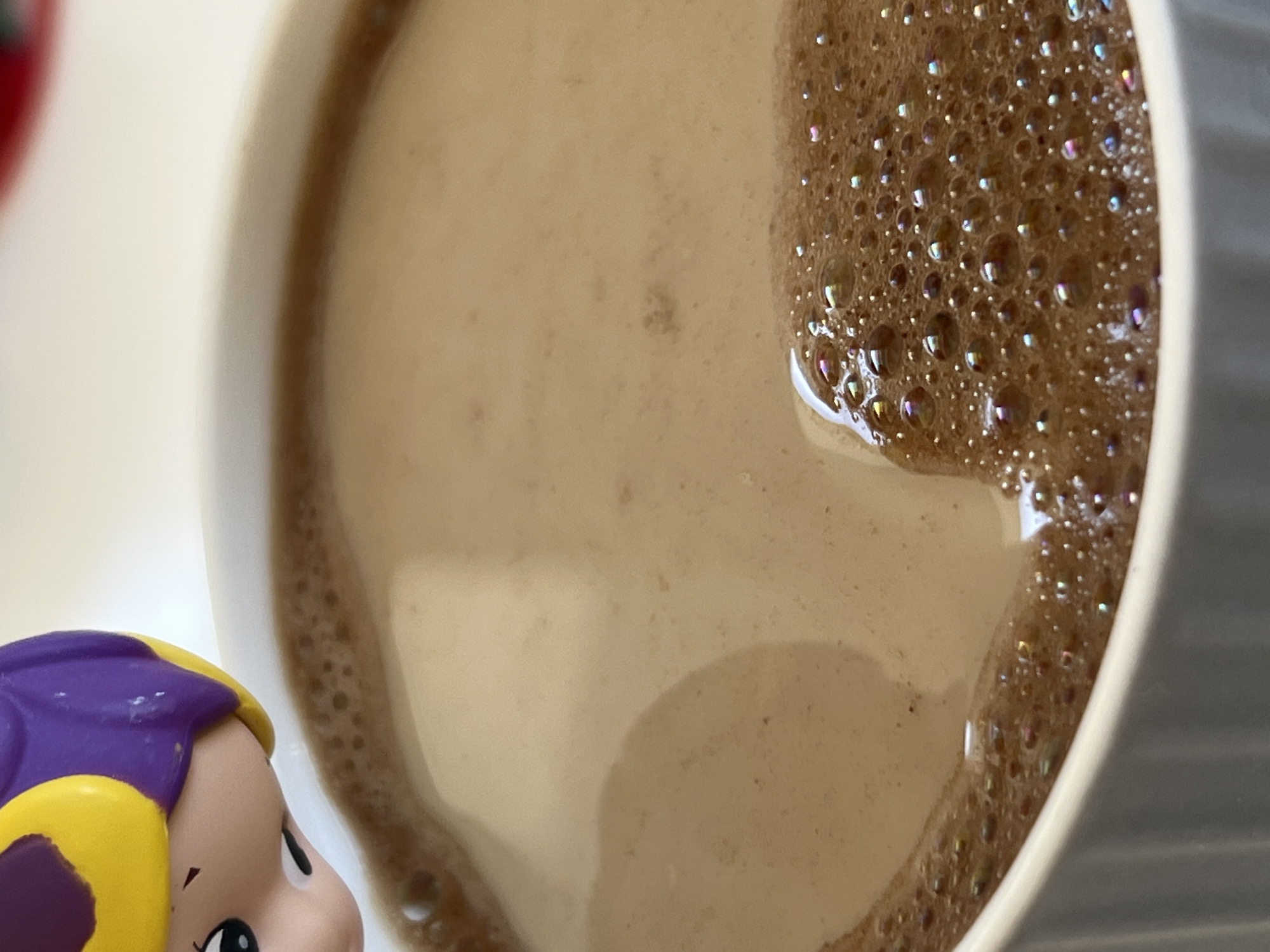 An overhead, close-up shot captures a creamy brown beverage in a white cup, with a small portion of a doll or toy visible in the bottom-left corner. The cup occupies most of the frame, showing a light brown liquid, likely coffee or hot chocolate. A thick layer of tiny, iridescent bubbles and foam covers the right side of the beverage and lines the inner edge of the cup. These bubbles reflect various subtle colors, suggesting light interaction.
The doll, or toy figure, has a light-skinned face, a dark eye looking upwards and to the right, and bright yellow hair partially covered by a purple accessory or hat. Only its head and a small part of its upper face are visible, positioned at the bottom-left edge of the frame, appearing to gaze towards the drink. The background is out of focus, primarily a soft white and light grey, suggesting an indoor setting. There is no visible text. The image presents a calm, possibly playful moment, with the doll serving as an unexpected companion to the beverage.