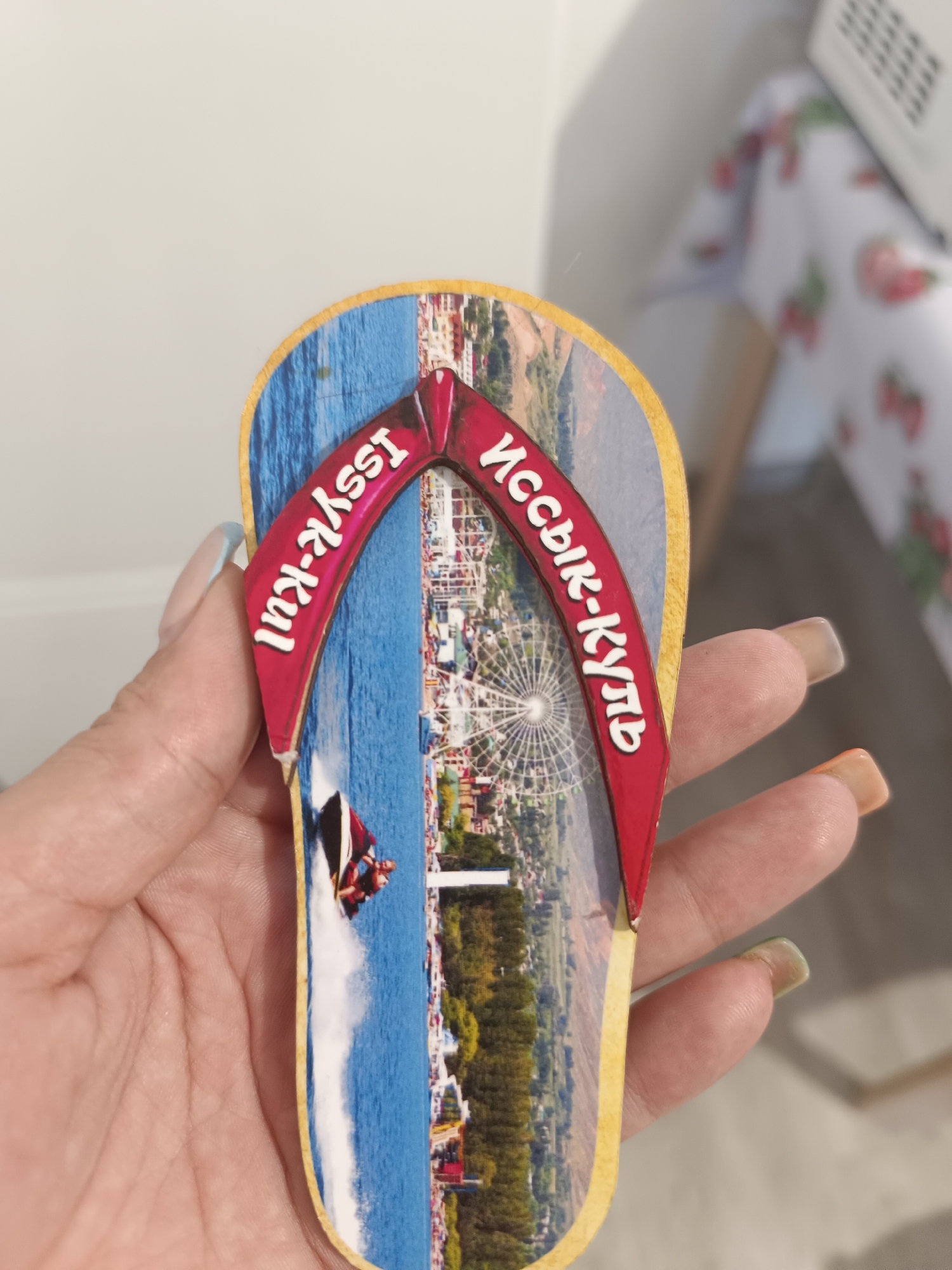 A close-up, overhead shot shows a hand with manicured nails holding a flip-flop shaped souvenir. The souvenir features a vibrant image of a jet ski in the water on one side and a Ferris wheel and cityscape on the other. Text in white cursive lettering appears on a red strap, with "Issyk-Kul" on the left side and what appears to be Cyrillic text on the right side. The background is out of focus, showing a white surface and a table with a strawberry-patterned tablecloth. The city administration of Taraz, Kazakhstan, is not depicted in this image.