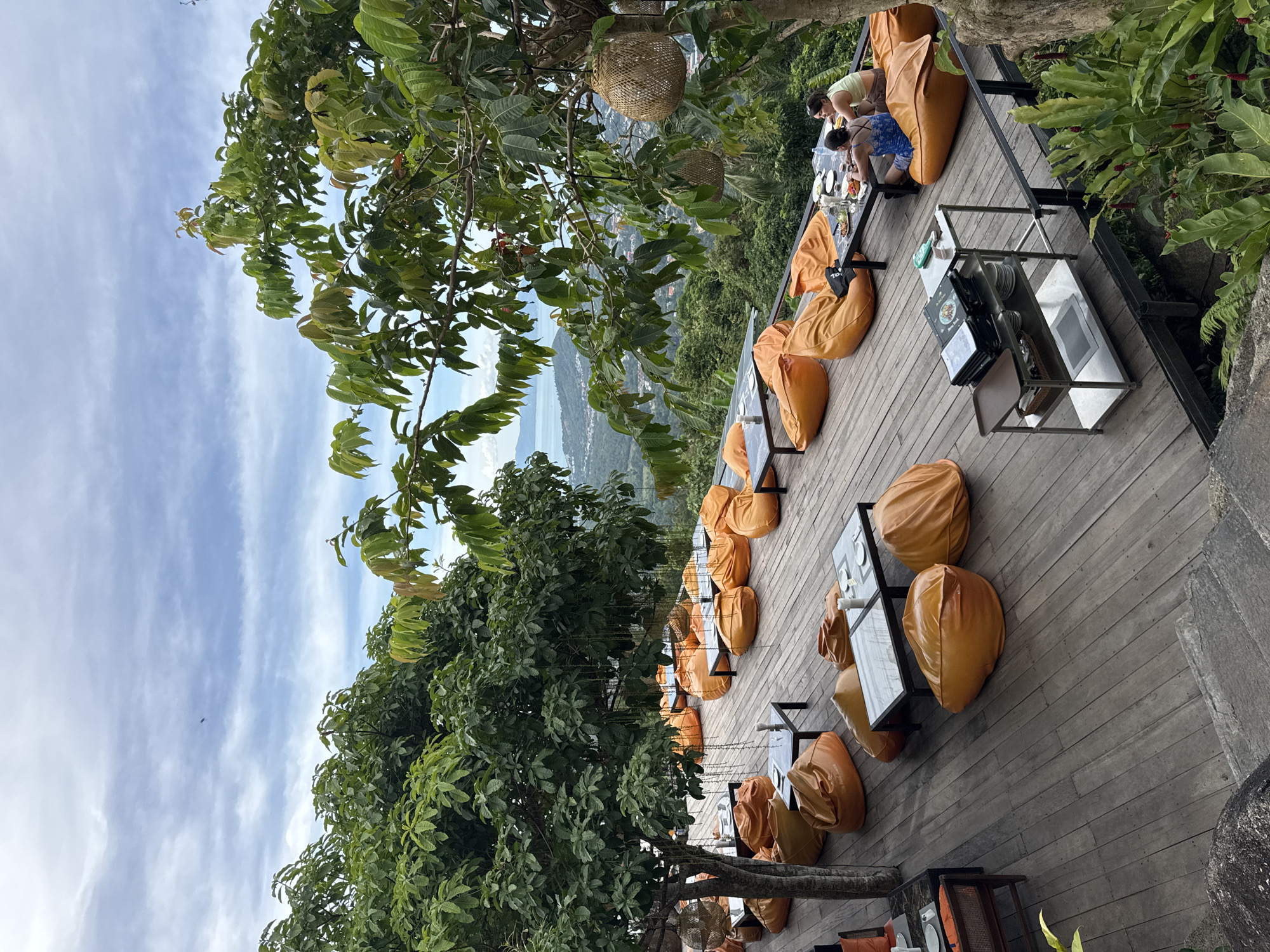 The image shows an outdoor seating area at a restaurant with a scenic view. The seating consists of numerous large, orange beanbag chairs and several low tables on a wooden deck. The tables are set with plates, utensils, and glasses. In the background, there is a lush green landscape with a body of water and buildings visible in the distance under a partly cloudy sky. Two women are seated at one of the tables, engaged in conversation and eating. There are large trees with green foliage providing shade. Some decorative wicker baskets are hanging from the trees. A metal serving cart with menus and dishes is positioned on the deck. The overall scene depicts a relaxed dining environment with a panoramic view.