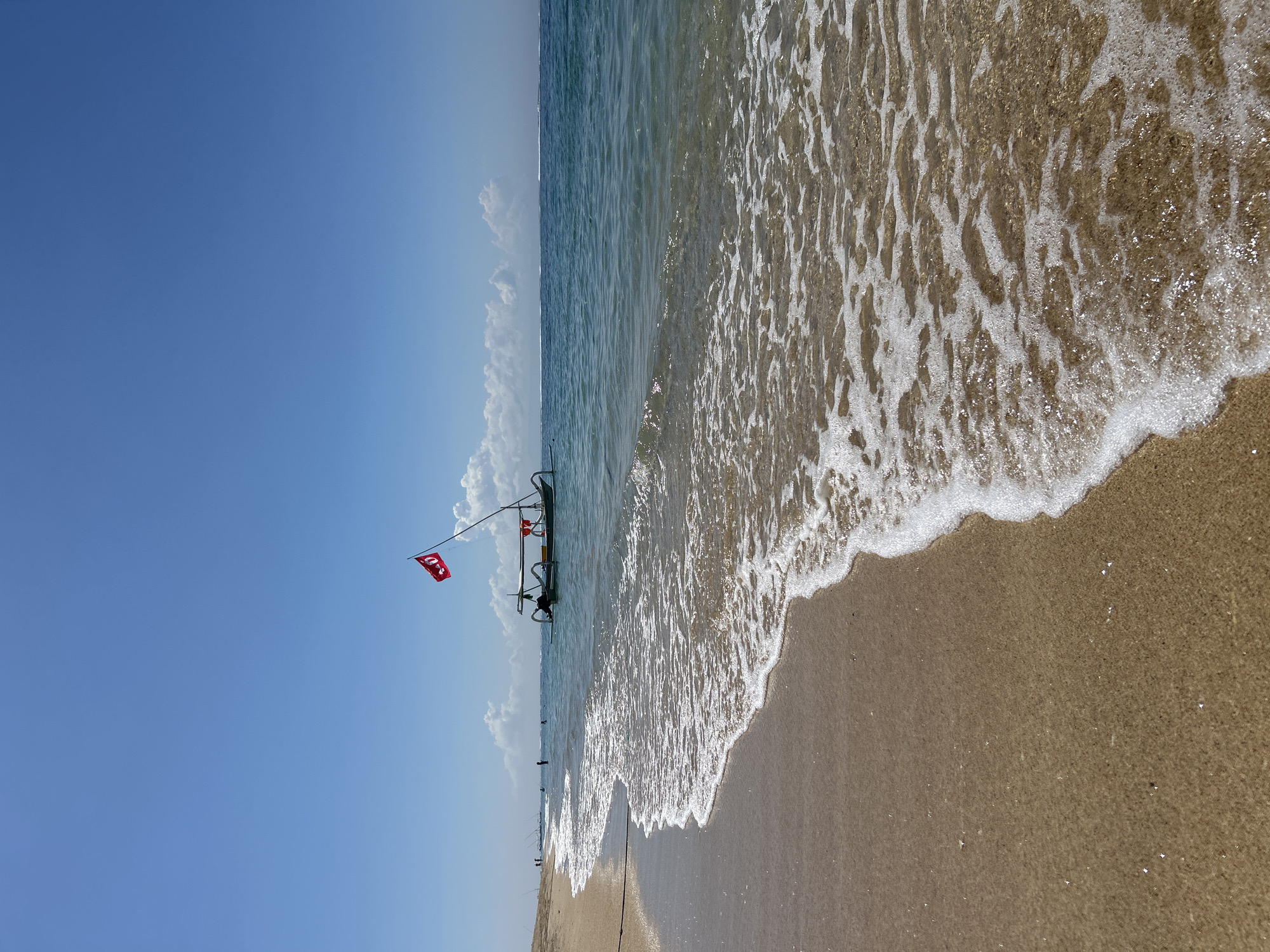 The image shows a beach scene, likely in Kuta Selatan, Indonesia. A lifeguard station with a red Turkish flag stands in the shallow water. The sky is clear blue with some clouds. Waves are washing onto the sandy beach. People are visible in the distance along the shoreline.