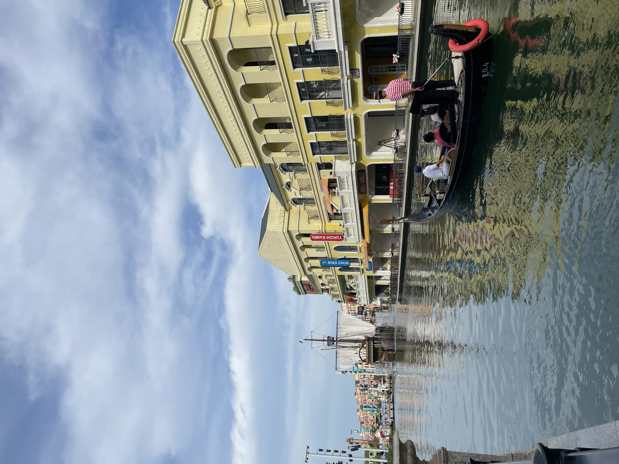 In the city of Nghĩa Trụ Commune, Vietnam, this image captures a vibrant canal scene with a Venetian theme. In the foreground, a black gondola, identified by the marking "E0.4", glides through the water. The gondola carries three individuals: a person in a white shirt and blue cap in the front, a person in a pink shirt in the middle, and a gondolier in a red and white striped shirt and a hat, standing and using a pole to propel the boat.
Along the right bank of the canal, a row of brightly colored, multi-story buildings with architectural elements reminiscent of Venetian design lines the waterway. These buildings feature arched windows, balconies with railings, and decorative cornices, painted in various shades of yellow, orange, and red. Some storefronts are visible at ground level.
In the middle ground, a large, wooden sailing ship with white sails is positioned in the canal, appearing to be part of a fountain display as water sprays from its sides. Further down the canal, a long line of similarly styled, brightly colored buildings stretch into the distance, creating a picturesque and lively atmosphere.
The sky above is a mix of blue with scattered white clouds. The water reflects the colors of the buildings and the sky, creating a shimmering effect on the surface. The overall impression is one of a man-made entertainment district designed to evoke the charm of Venice.