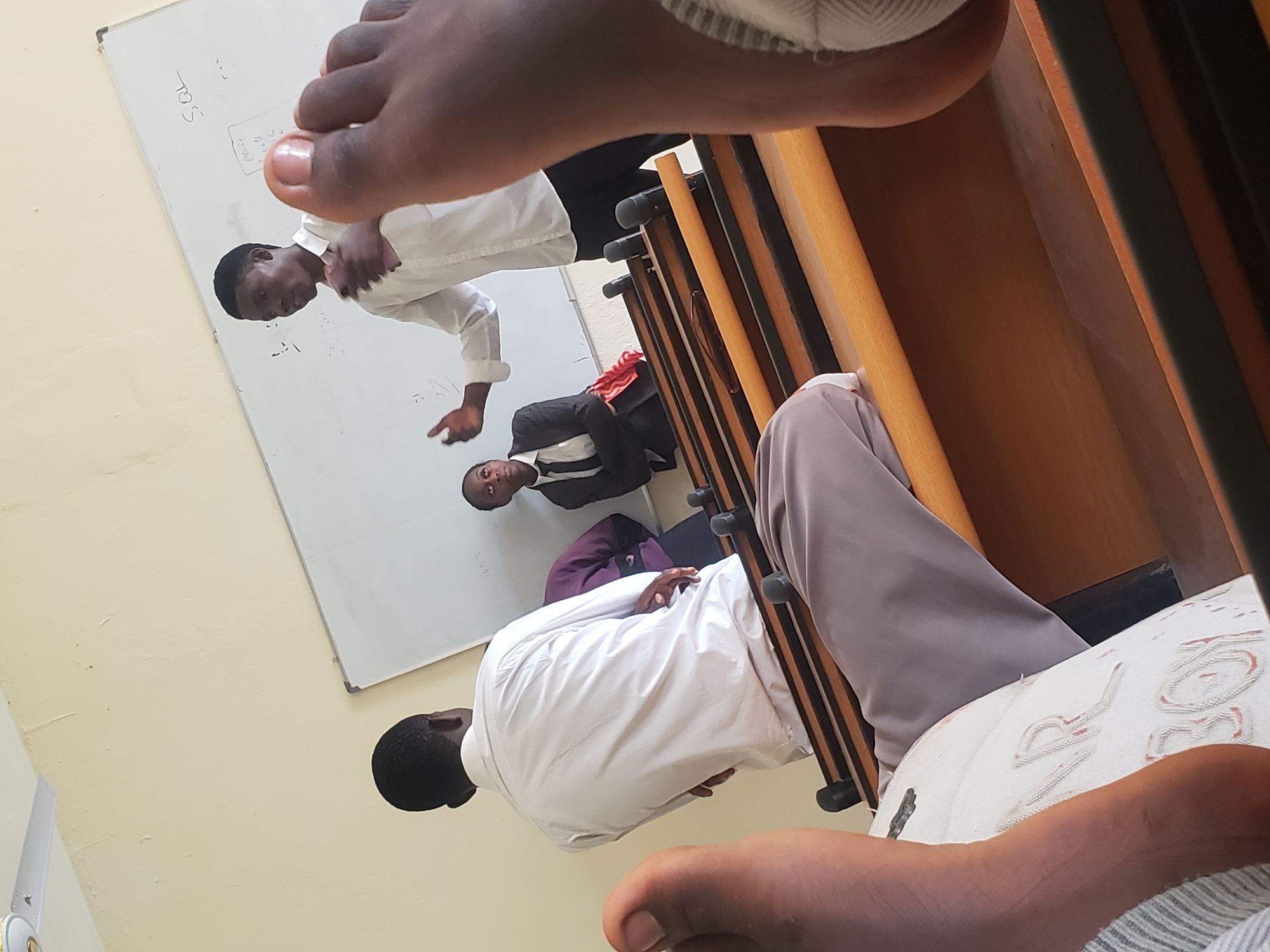 In a classroom setting in Gumel, Nigeria, a lecture is in progress. A young man in a white shirt stands near a whiteboard, gesturing with his hands as he speaks. To his left, another young man in a white shirt faces away from the camera, seemingly engaged in the lecture. To the right of the presenter, a young woman in a black suit and tie looks up towards the whiteboard. In the foreground, the perspective is from someone lying down, with their feet visible in the lower portion of the frame, resting on a white surface with lettering. Wooden tiered seating characteristic of a classroom or lecture hall is present. The whiteboard has some writing on it, including "SQL."