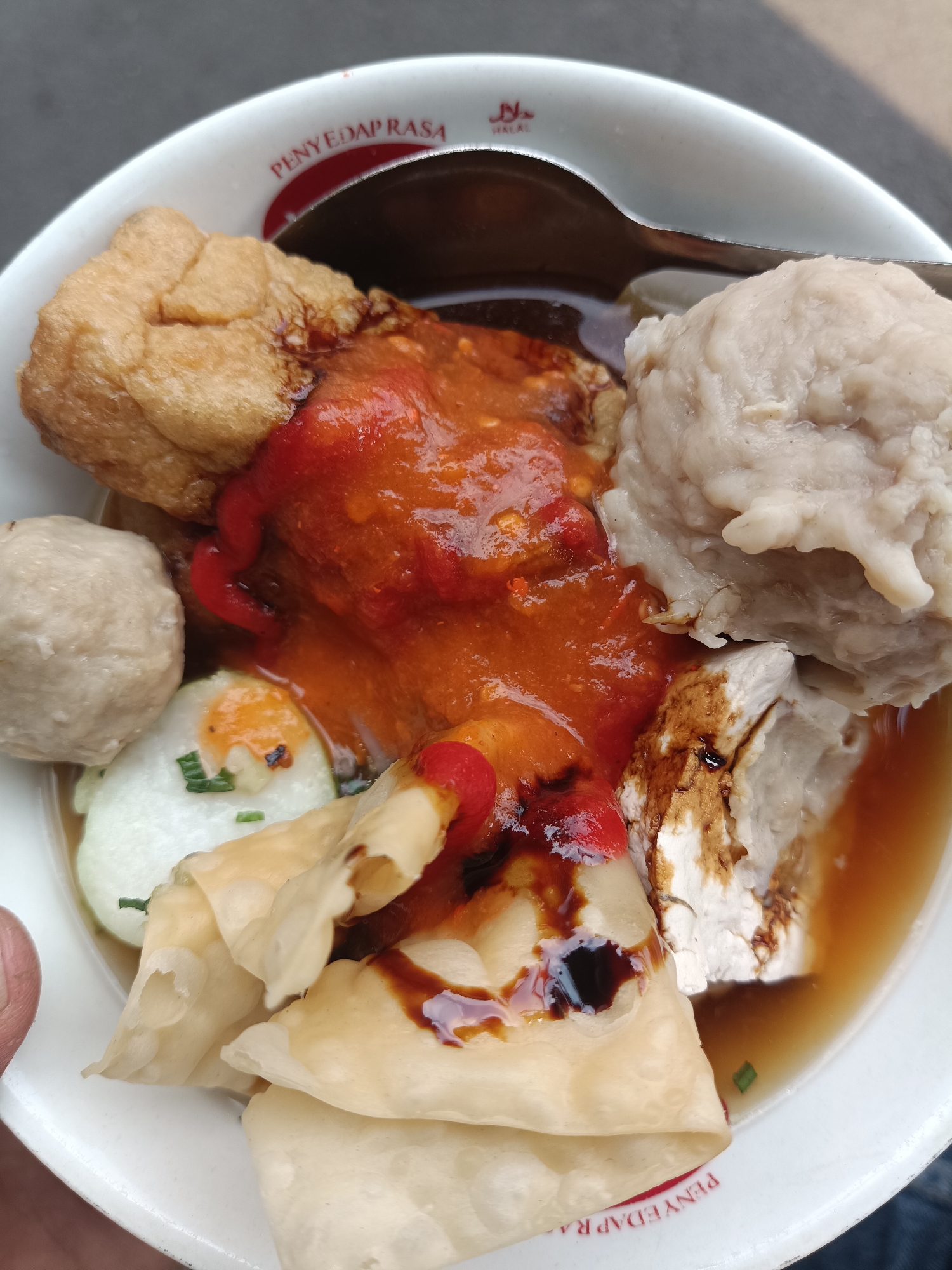 This is a close-up, eye-level, overhead shot of a bowl of Indonesian meatballs, known as bakso, served in a white ceramic bowl with red lettering on the rim that reads "PENYEDAP RASA" and a halal logo. The bowl contains several meatballs, some whole and some cut. One large meatball is prominent in the upper right, with another smaller one to its left. There are also pieces of fried tofu, a sliced hard-boiled egg with chives, and folded pieces of what appears to be wonton skin or a similar dough wrapper. The bakso is covered in a vibrant red chili sauce and drizzled with dark soy sauce or a similar condiment. The broth is a light brown color. A spoon is partially visible in the upper portion of the bowl, resting against the rim. The background is a textured gray surface, likely a table. The image does not provide specific details about the city of Jember, Indonesia, other than the possibility that "PENYEDAP RASA" may be a local restaurant name or a common food seasoning brand in the region.