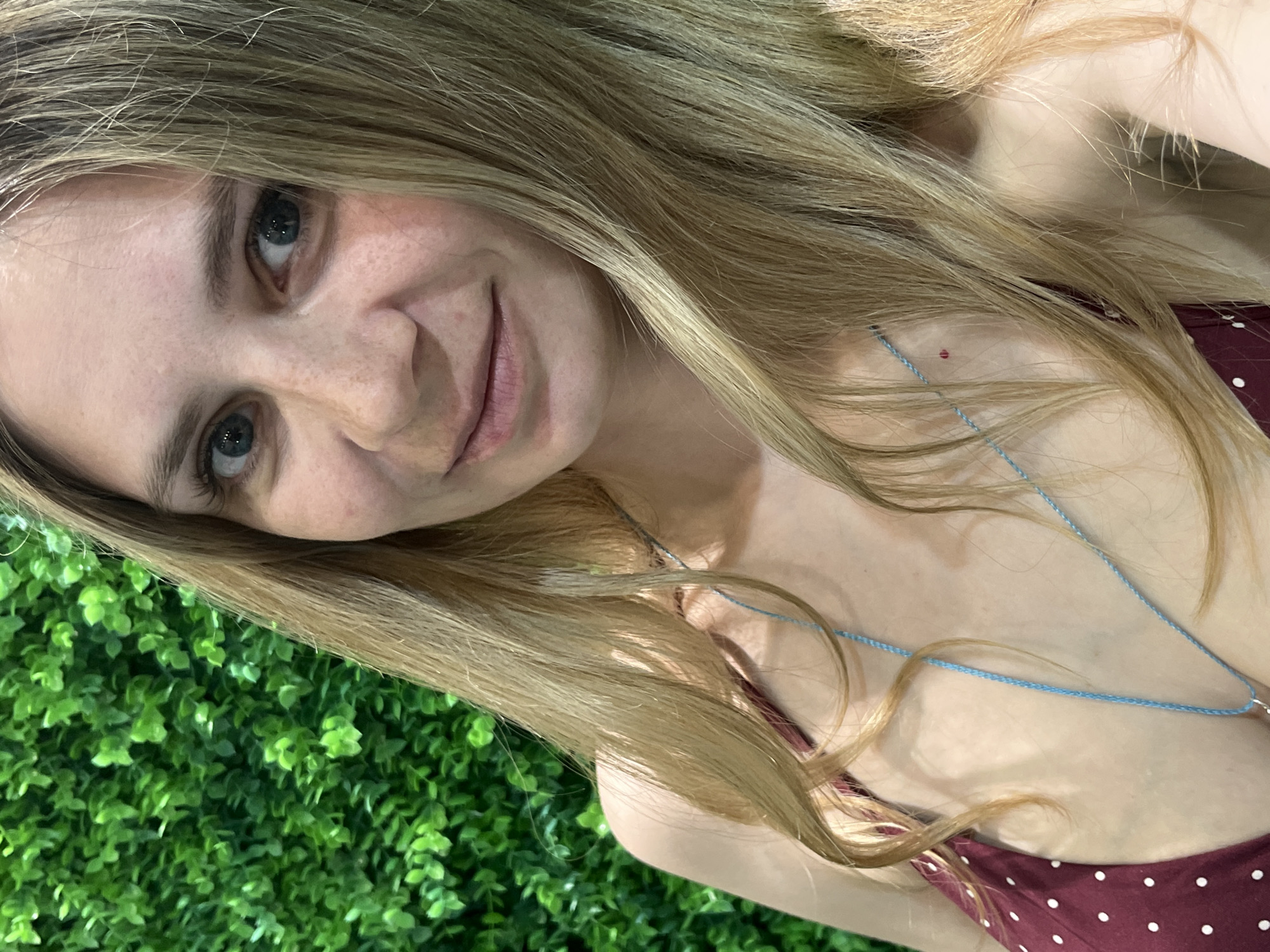 A young woman with blonde hair and blue eyes poses for a selfie. She is wearing a red and white polka dot top with a thin blue necklace. The background features a green artificial plant wall. It is impossible to ascertain the location in the city of Chaikovsky, Russia, from this image.