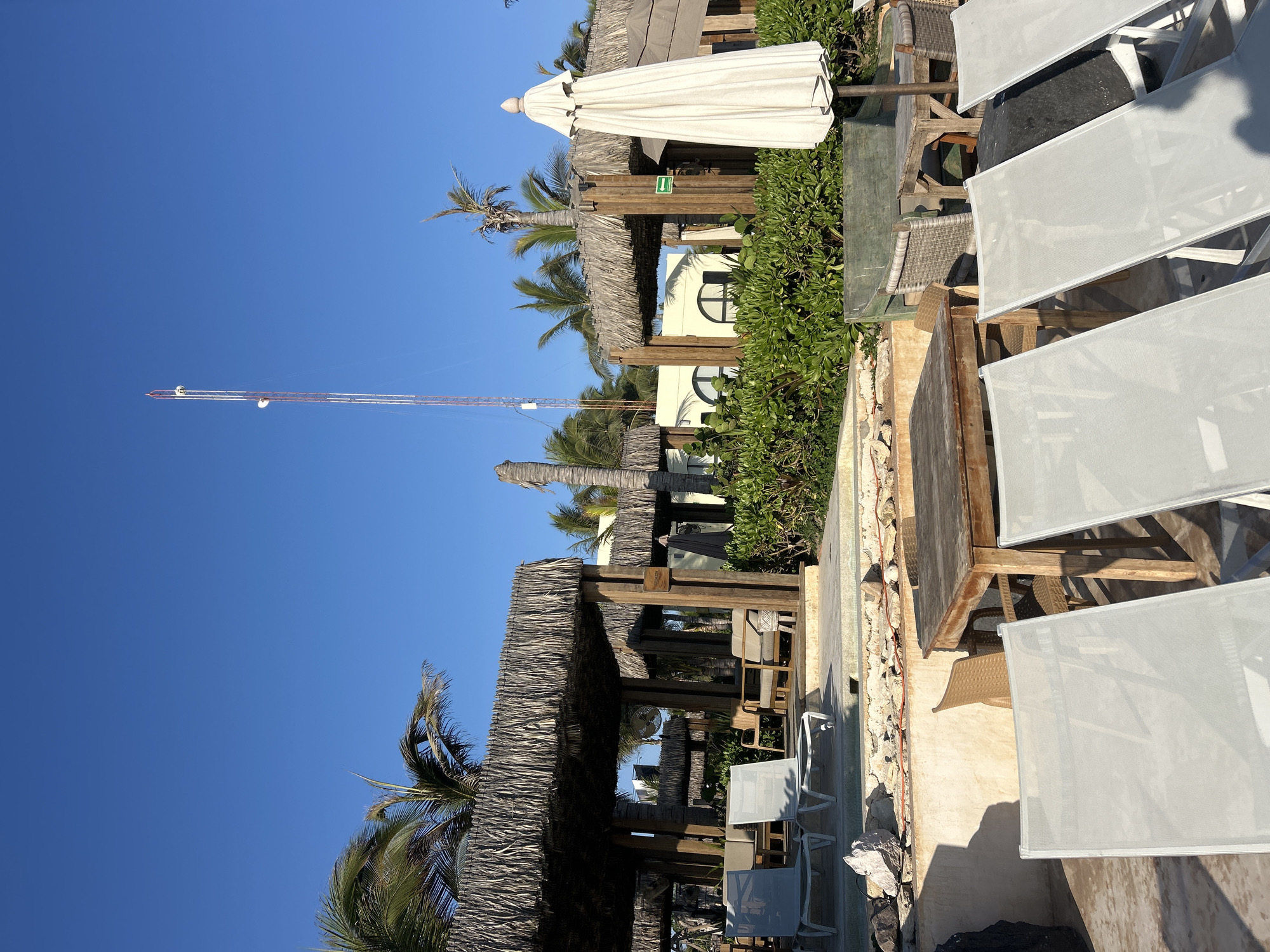 This image depicts a sunny outdoor area, likely part of a resort or beachfront establishment, under a clear blue sky in Mazatlán, Mexico. The bright sunlight suggests it's midday or early afternoon, casting distinct shadows across the scene.
In the foreground and extending into the mid-ground on the right, several empty lounge chairs with light grey fabric are arranged on a paved patio or deck. To the left of these chairs, a walkway leads to a series of shaded structures. These structures feature traditional thatched roofs, known as palapas, supported by robust wooden posts, providing a tropical ambiance. Underneath the palapas, wooden tables and chairs are arranged for outdoor dining or relaxation. A large, closed white umbrella stands near one of the shaded areas.
Lush green foliage, including manicured bushes and several tall palm trees, is interspersed throughout the scene, contributing to the resort-like setting. In the background, a light-colored building with distinctive arched windows is visible, likely part of the main complex. A small green sign with a white arrow pointing to the right is affixed to one of the wooden posts within the shaded area. On the far left, a tall, slender structure, possibly a communication tower or a distant crane, extends high into the clear sky. The area appears quiet and ready for guests, with no people visible in the frame.
