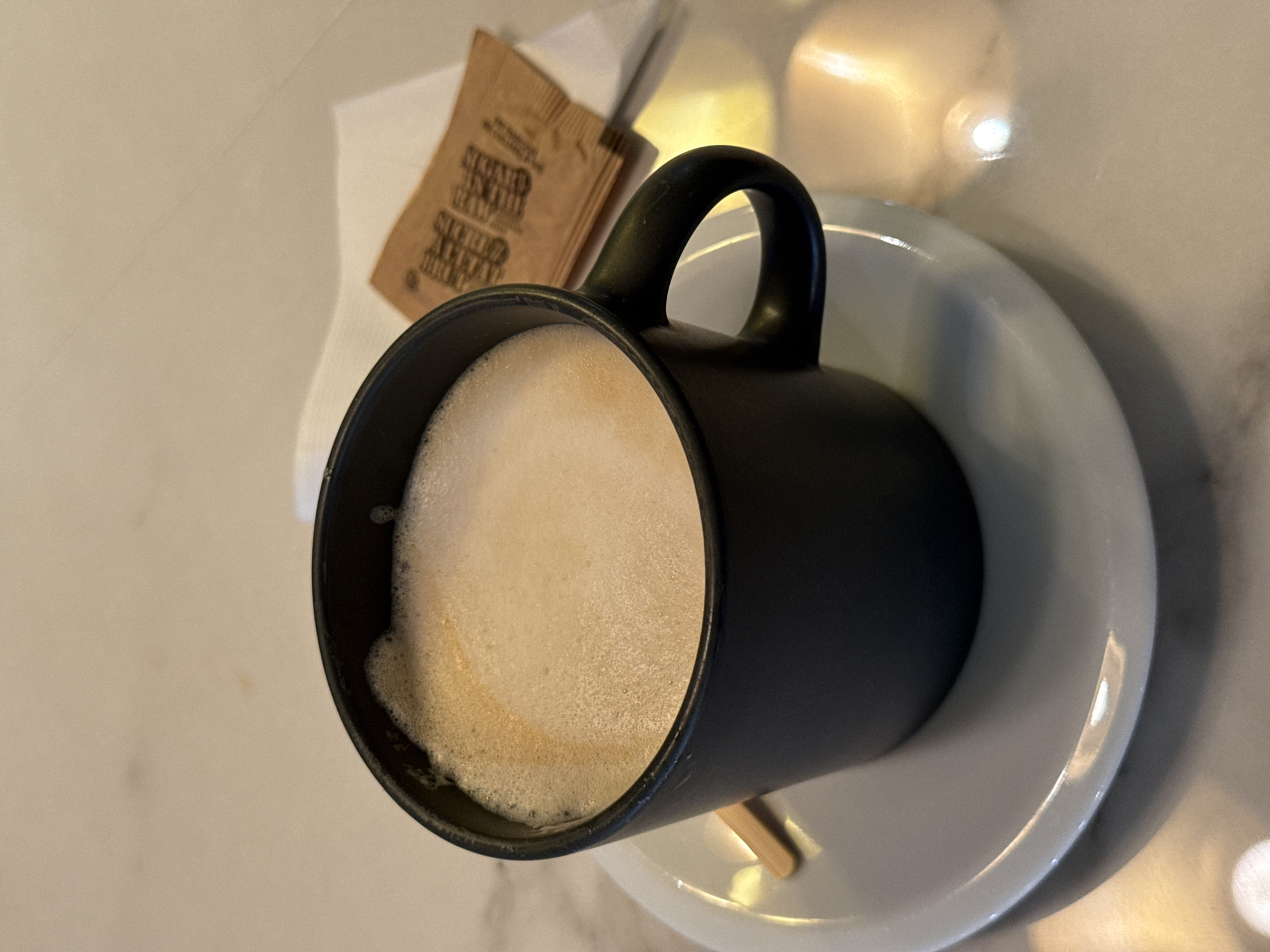 The image presents an overhead, slightly angled view of a black ceramic mug filled with a frothy, light-brown beverage, likely coffee or a latte. The mug rests on a simple white saucer, which in turn is placed on a light-colored, reflective tabletop, possibly made of marble or polished stone. A small, light-colored wooden stirring stick is visible on the saucer, partially obscured by the mug.
Behind the mug, two small brown packets are neatly arranged on a white paper napkin. These packets appear to contain sweeteners. Prominently visible text on the packets is in French, reading "SUCRERIE DE LA GUEULE" and "NATURELLEMENT SUCRÉ AU SIROP D'ÉRABLE." These phrases translate to "Sweet treat for the mouth" and "Naturally sweetened with maple syrup," respectively. The mention of maple syrup serves as a distinct cultural and geographical indicator, aligning with the provided context that the image is from Montreal, Canada.
The lighting in the scene suggests an indoor environment, possibly a cafe or lounge. Reflections of light sources create bright highlights on the tabletop and saucer, indicating a smooth, glossy surface. Shadows are cast by the mug and saucer, adding depth to the composition. The overall arrangement suggests a ready-to-be-consumed beverage service. No people are visible, and the scene is calm and focused on the objects.