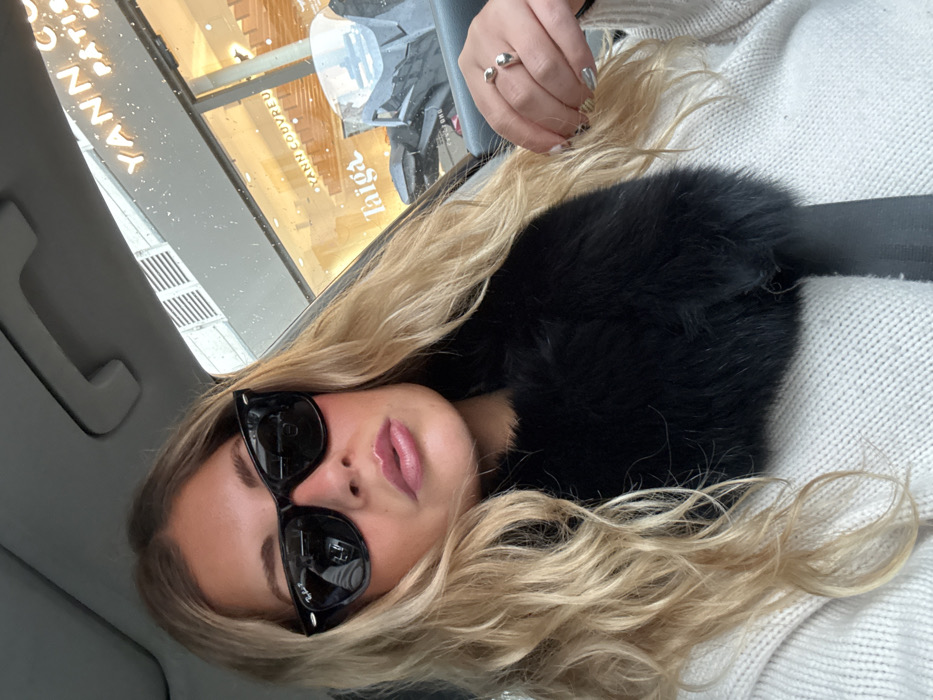 A blonde female is seated in the front passenger seat of a vehicle, looking directly at the camera. She wears black Ray-Ban sunglasses, a cream-colored chunky knit sweater, and a black fur collar or scarf. A black seatbelt is fastened across her chest. Her right hand, adorned with a silver-colored ring with two spherical elements, is visible near her shoulder. Through the wet vehicle window, the illuminated storefront of "YANN COUVREU PÂTISSIER" and "Taiga" is visible, indicating the location is in Paris, France. The exterior building facade is light-colored with horizontal louvered blinds on an upper floor. The overall scene suggests wet weather conditions.
