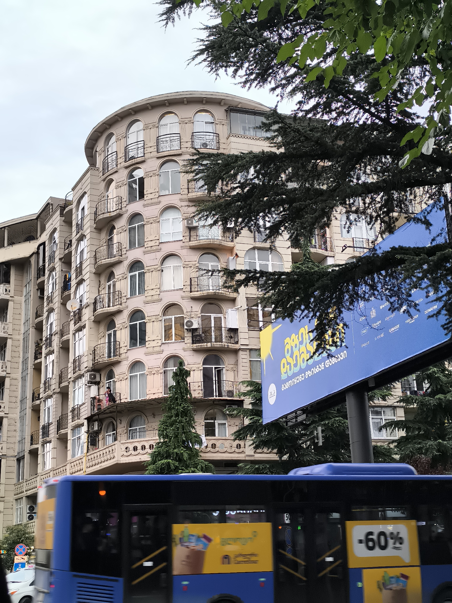 A view of a building in Batumi, Georgia, with a blue bus passing in the foreground. The building, likely an apartment complex, is light pink with a curved facade. It features numerous arched windows, balconies with decorative railings, and air conditioning units. Some windows are open, and a few people can be seen on balconies. A tall evergreen tree stands in front of the building. To the right of the building is a large blue billboard with text in Georgian and a yellow graphic. A portion of a blue bus, with yellow advertisements on its side showing discount percentages and product images, is blurred due to motion in the foreground. A white car and traffic signs are visible to the left of the bus. The sky is overcast.