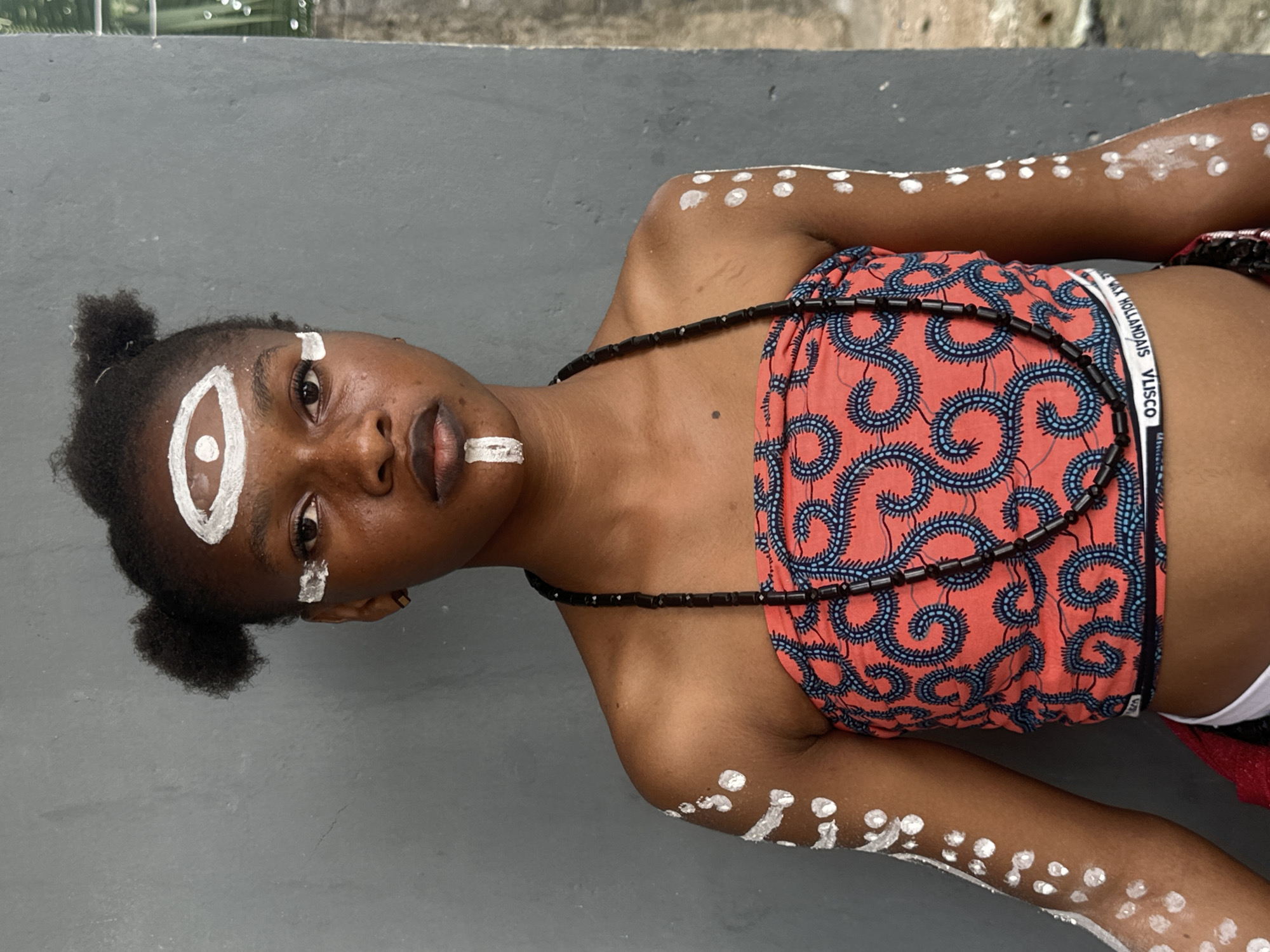 A young Black female, likely a teenager, stands facing the camera against a muted gray wall. She has dark skin and her hair is styled in two small buns on top of her head. Her face is adorned with white paint: a large, almond-shaped design surrounds her forehead and eyes, with a smaller dot in the center representing an eye. A vertical white line is painted on her upper lip. She wears a strapless, coral-colored top with a swirling blue pattern, and a black beaded necklace. Her arms and shoulders are decorated with white polka dots and streaks. The background is a plain, textured gray wall, with a sliver of greenery visible on the right side, suggesting an outdoor or semi-outdoor setting. The lighting is natural and appears to be overcast, creating soft shadows. The overall impression is one of cultural or ceremonial expression. The image is set in Badagry, Nigeria, a historic coastal town known for its rich cultural heritage and slave trade history.