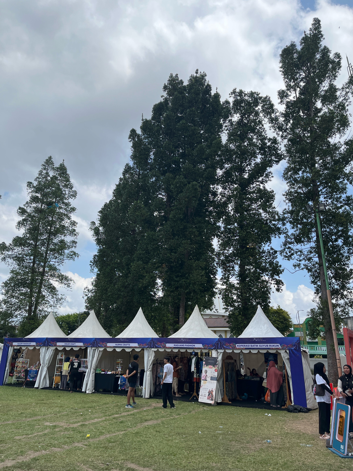 The image shows an outdoor market with several tents set up on a grassy field. The tents are white and peaked, and some have blue banners with text on them. People are seen browsing the stalls and interacting with each other. The stalls appear to be selling various goods, including clothing and crafts. Tall trees are visible in the background, and the sky is partly cloudy. The scene is likely taking place in Lumajang, Indonesia, given the context.