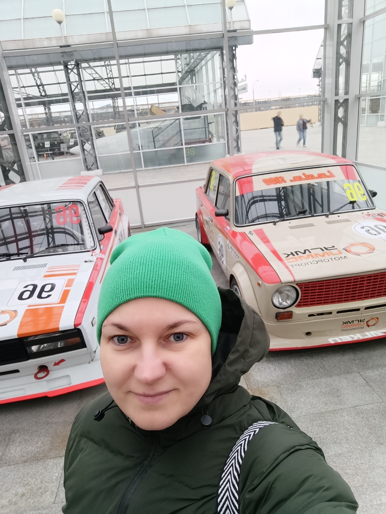 A woman takes a selfie indoors, with two vintage-style race cars behind her. She stands slightly off-center to the left, facing the camera with a neutral to slight smile. She has fair skin, light-colored eyes, and wears a bright green knit beanie and a dark green puffer jacket.
The two race cars are positioned to her right and slightly behind her. The car on the left is primarily white with prominent orange and red stripes and accents. Large black letters "ae" are visible on its hood, and a red number "86" is on its side window. A red tow hook is present on its front bumper. The car on the right is predominantly beige with similar red and orange racing stripes and details. It features a red grille. Yellow number "88" is displayed on its windshield sun strip. On its side, there's a white circle with an orange border containing "ae", and below it, a white number "38" (or "88") with an orange outline. Mirrored text on its front bumper/valance and hood appears to read "ALMK" and "МОТОСПОРТ" (Motosport).
The setting appears to be a modern indoor space, possibly a museum or exhibition hall, characterized by large glass panels and a metallic structural framework. The floor is light-colored pavement or tiling. Through the glass, an overcast daytime sky and two blurred figures walking in the distance are visible, along with glimpses of other buildings or structures. The scene captures the woman interacting with an automotive display, likely during a visit to an exhibition in Verkhnyaya Pyshma, Russia.