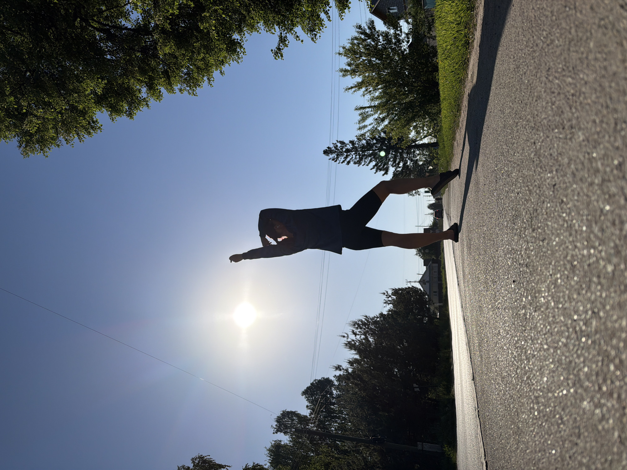 A low-angle, outdoor daytime shot shows a person in silhouette in Chaikovsky, Russia, against a bright sky and a sunburst. The person is wearing dark clothing, including what appears to be a long-sleeved shirt and shorts, and is positioned horizontally as if "flying" or leaping. The backdrop is comprised of the sky, some trees and foliage, and a portion of a paved surface. Power lines are visible against the sky. A building and some grass are visible in the upper right.