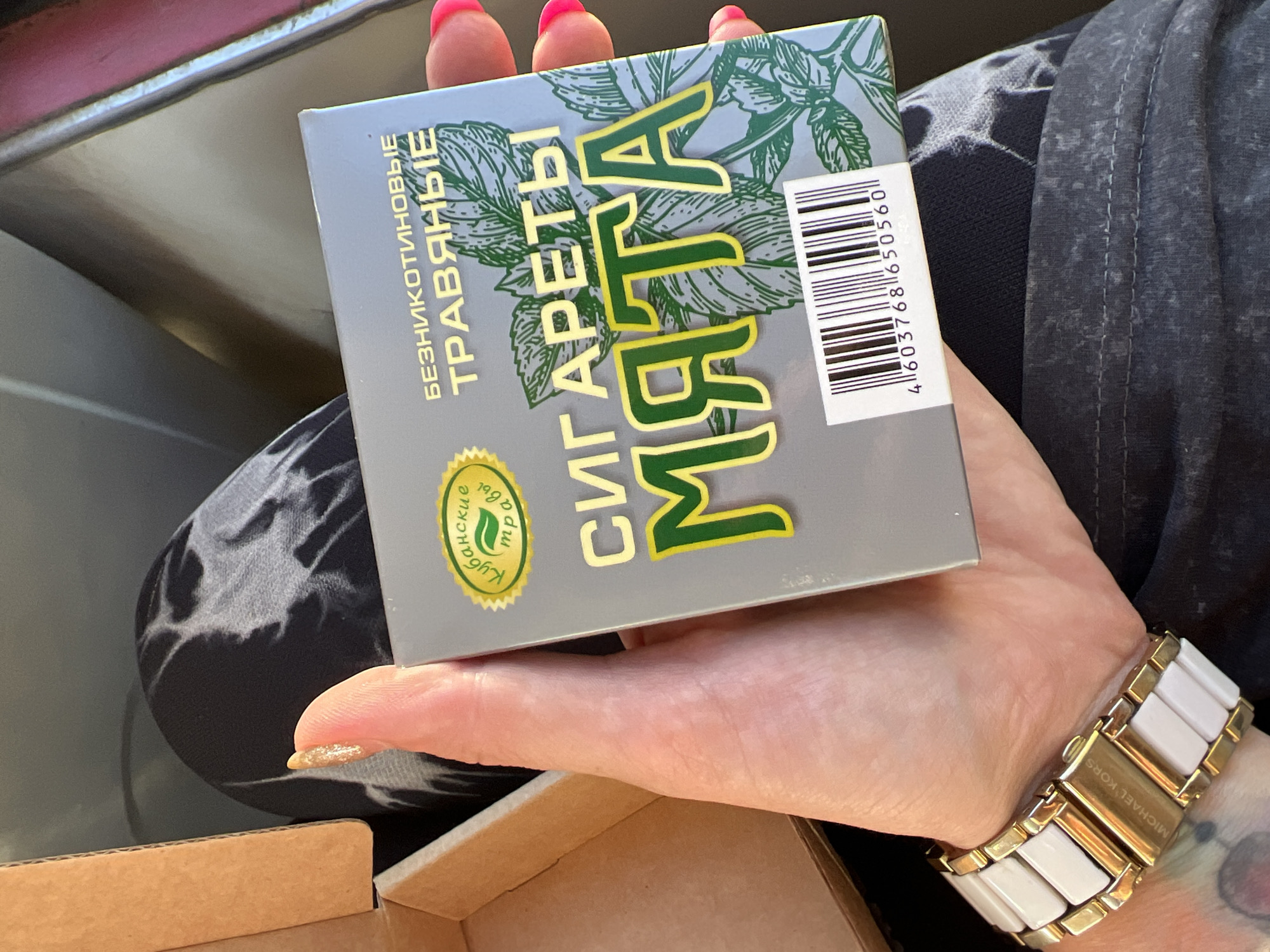 A close-up, waist-up shot captures a person holding a rectangular box of herbal cigarettes in their left hand. The box is predominantly gray with green and yellow text and imagery of mint leaves. Prominently displayed on the box are the words "СИГАРЕТЫ МЯТА" in large, green, outlined letters. The person's hand, with bright pink acrylic nails, is holding the box from the side, with fingers curled around it. A gold and white ceramic watch is visible on the person's wrist. The background consists of dark, textured fabric, possibly leggings or a seat, with a glimpse of a cardboard box on the left side of the frame. The scene appears to be indoors.