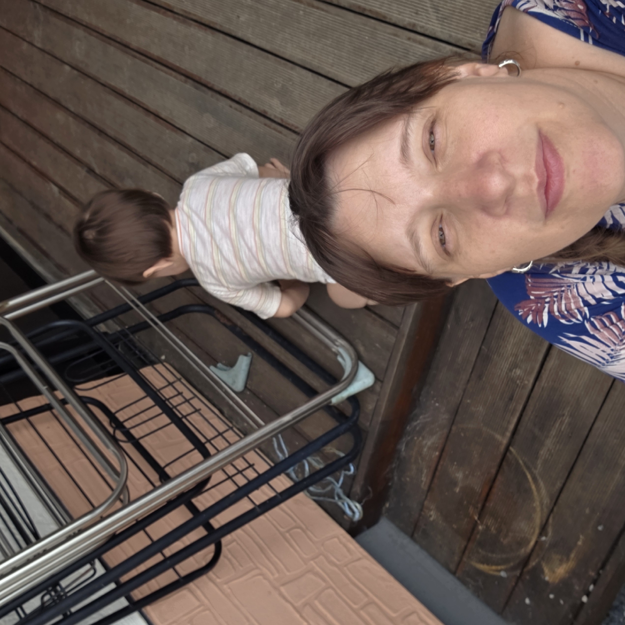 The image shows a woman and a child on a wooden deck. The woman is in the foreground, taking a selfie, and the child is in the background, facing away from the camera. The child is wearing a striped shirt and appears to be crouching. There is a metal drying rack and a pink brick wall visible behind the child. The deck is made of wooden planks, and there are some dirt and scuff marks on the surface. The scene is outdoors, and the lighting suggests it is daytime. The location is stated to be Dunnae-myeon, South Korea, which is likely a rural or suburban area given the outdoor setting.