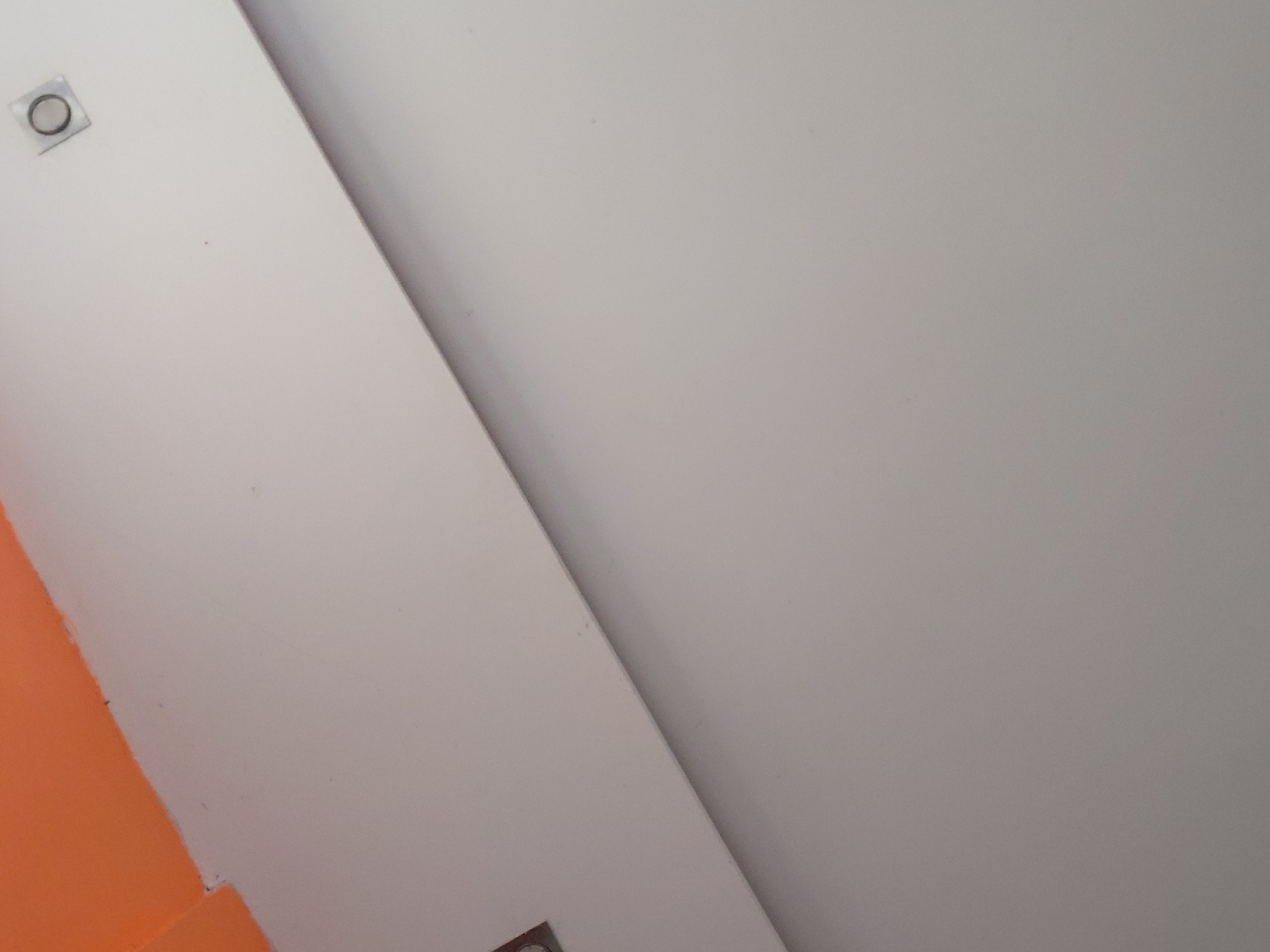 This image presents an abstract, architectural view of an interior space, likely within a building in Santa Ana, Venezuela. The composition is characterized by large, intersecting planes of color and texture.
A dominant diagonal line slices across the frame, dividing a brighter, off-white surface on the left from a slightly darker, light grey surface on the right. This demarcation suggests an interior corner, a seam between panels, or a change in material or lighting.
In the upper-left quadrant, embedded within the off-white surface, is a small, circular light fixture. It features a metallic-looking square bezel encircling a dark, recessed circular element. Another similar fixture is partially visible at the bottom edge of the white surface, towards the center-right.
A striking, solid orange surface occupies the lower-left portion of the image. It forms a sharp, angular boundary with the off-white surface, suggesting it is a wall or a structural element contrasting boldly with the muted tones of the rest of the space.
All visible surfaces appear smooth and clean, with a subtle, uniform texture, likely from paint or plaster. The lighting is even and diffused, creating no harsh shadows. There are no people, objects (beyond the light fixtures and architectural elements), or activities depicted. No visible text is present. The overall impression is one of modern, minimalist interior design.