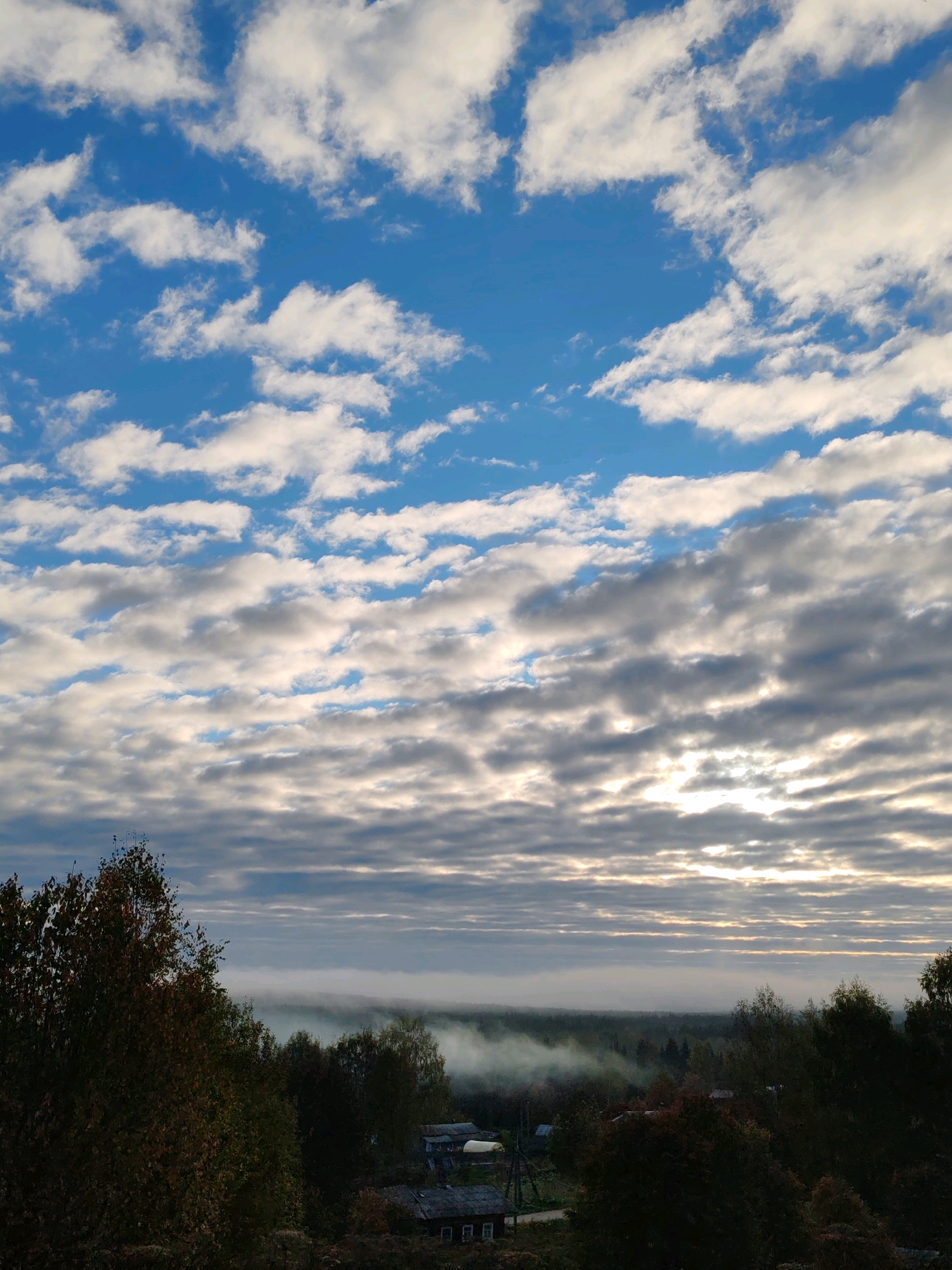 The image depicts a rural landscape at dawn or dusk, with a vast expanse of sky filled with layered clouds. The lower portion of the image reveals a village scene in Chukhlem, Russia, characterized by several houses with dark roofs. A gentle mist or fog hovers over the distant treeline, suggesting a cool or damp atmosphere. The foreground is dominated by the dark silhouettes of deciduous trees, some showing hints of autumn foliage. The overall lighting is subdued, with the sun's rays partially breaking through the clouds, casting a soft glow on the scene. There are no people visible in the image.