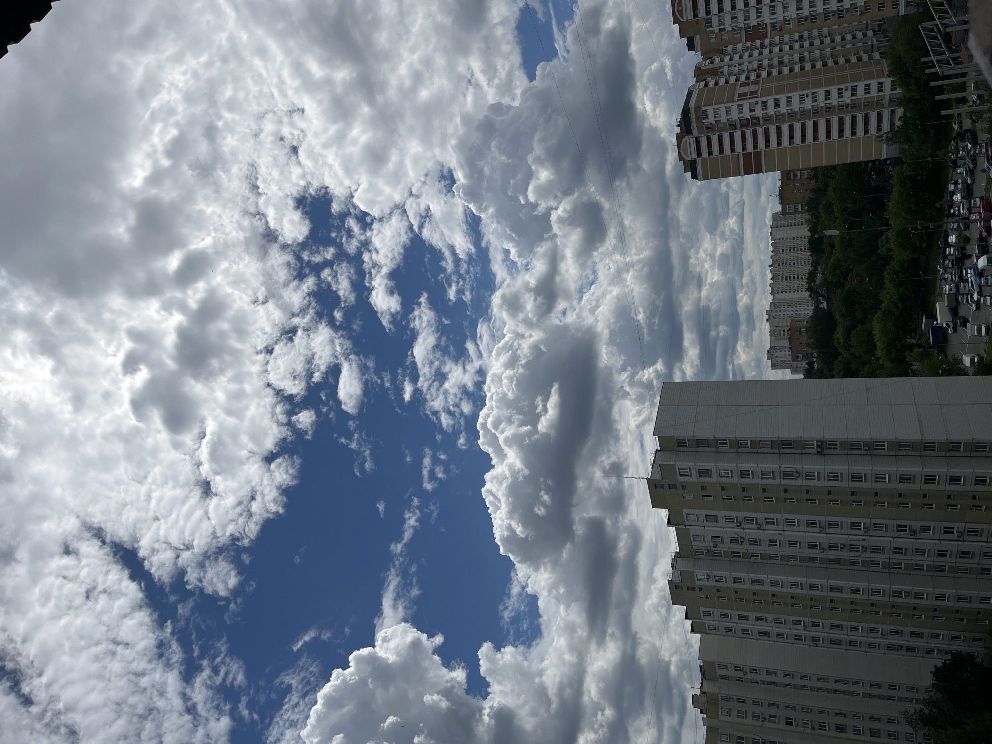 The image depicts a view of a residential area in Moscow, Russia, under a partly cloudy sky. The foreground features several tall, multi-story apartment buildings with a light-colored facade and numerous windows. These buildings are characteristic of Soviet-era mass housing. In the middle ground, a dense line of green trees separates the apartment buildings from a parking lot filled with a variety of cars. The background shows more apartment buildings, also of similar architectural style, stretching into the distance. The sky is a vibrant blue in some areas, with large, fluffy white and gray clouds dominating the upper portion of the frame, suggesting dynamic weather conditions. Power lines are visible crisscrossing the sky, a common feature in urban landscapes. The overall impression is of a typical urban environment on a day with active cloud formations.