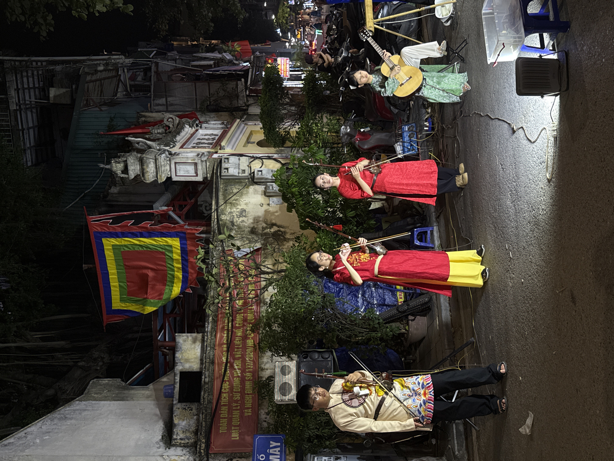 A street scene at night in Hà Nội, Vietnam, shows four musicians performing. One woman plays a guitar while reclining on what appears to be parked motorbikes. Three other musicians stand in a row, each holding and playing a stringed instrument, possibly a type of fiddle. The two women in the middle wear red robes with yellow accents, while the musician at the front wears a light-colored top with a decorated vest over black pants. A banner with a geometric pattern hangs on a nearby building. A sign with Vietnamese text is visible near the bottom left of the frame, and parked motorbikes and lights from businesses are visible in the background. A small speaker and light are present next to the musicians.