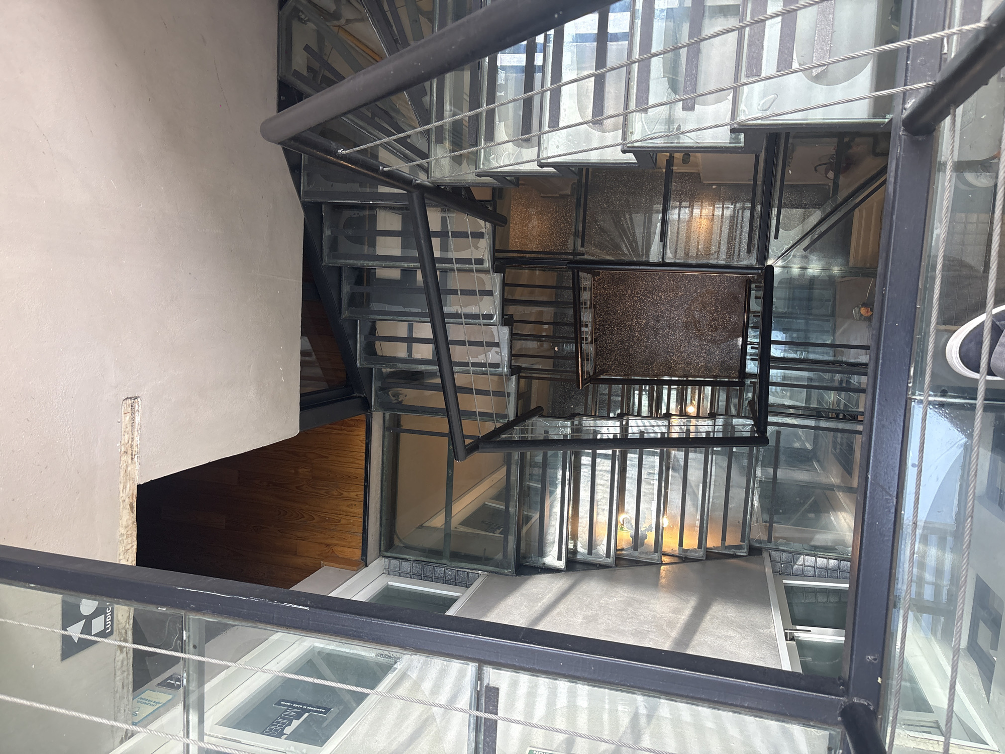 The image presents a bird's-eye view of a spiral staircase in what appears to be an interior space, with a section revealing a wooden floor. The staircase's design is modern, featuring glass treads framed by a dark metal structure, possibly steel or iron. A rope cable runs alongside the staircase as part of the safety railing.
The presence of advertising signage, only partially visible, and the general architectural style suggests the location is likely in an urban setting. Considering the "MUJERES" and "LUDIC" signs, and the context of a modern architectural staircase design, the image may have been taken inside the Soumaya Museum in Mexico City, Mexico.
There is one person visible, with only their foot and shoe visible on the right side of the picture.