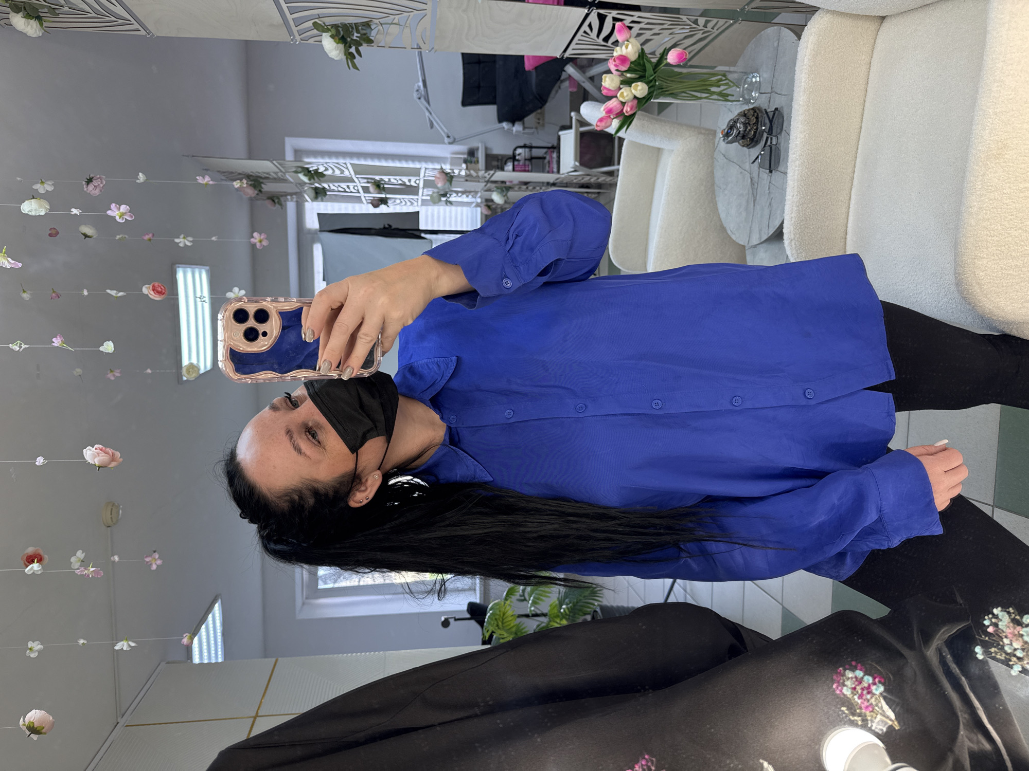 A woman wearing a black face mask and a bright blue button-down shirt is taking a selfie in a mirror. She is holding a cell phone with a decorative case. Her long black hair is visible. The reflection shows a well-lit interior space with decorative floral arrangements hanging from the ceiling. There are also chairs and a small table with flowers in the background. The setting appears to be a salon or boutique. The location is in Chaikovsky, Russia.