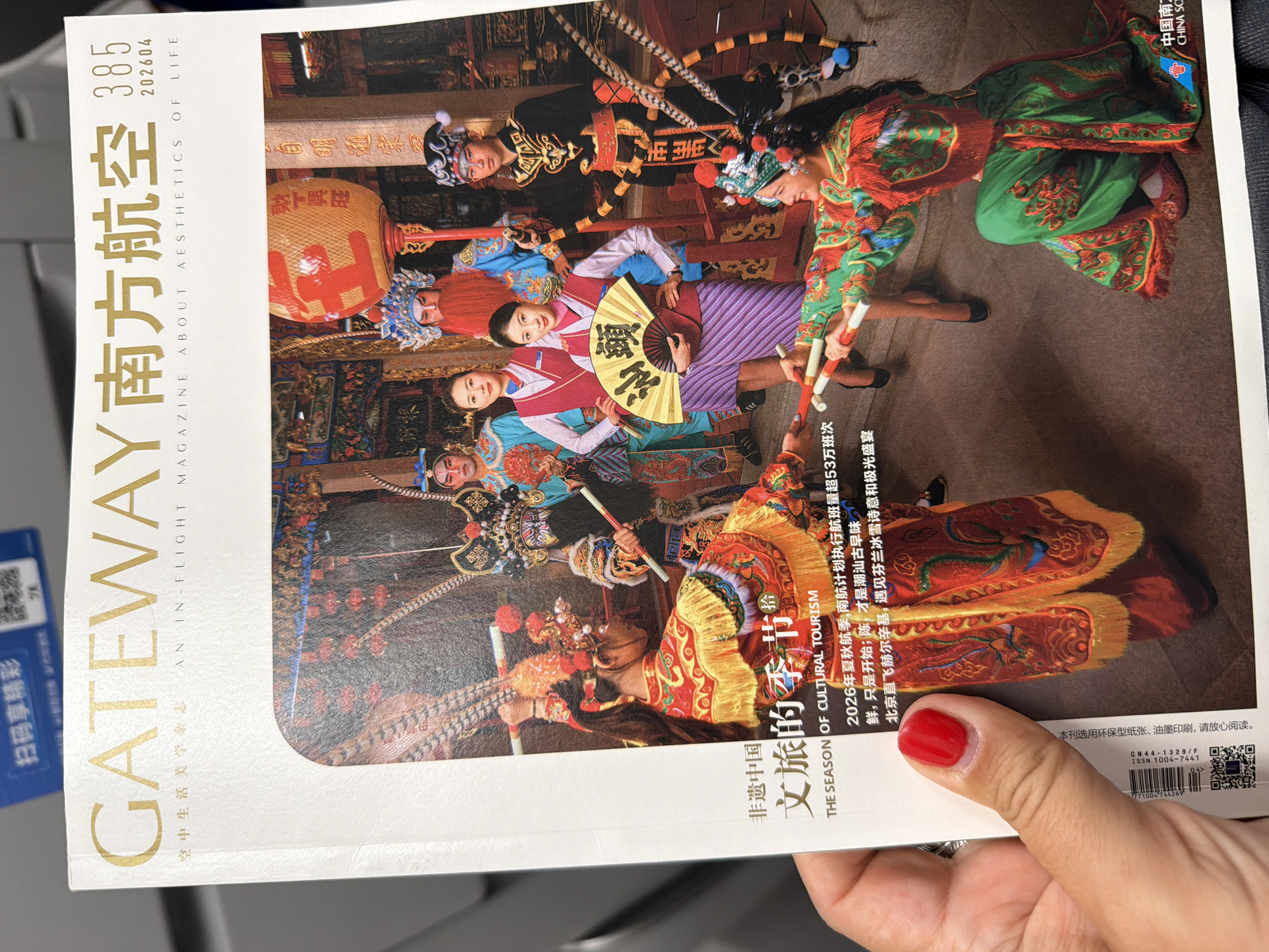 The image displays the vibrant cover of the "GATEWAY 南方航空" (China Southern Airlines) in-flight magazine, issue 385, dated 202604. The cover's central theme, highlighted by the title "非遗中国 文旅的季节" (Intangible Cultural Heritage China, The Season of Cultural Tourism), features a dynamic tableau of Chinese traditional performance.
The main subjects are several individuals, likely performers, adorned in elaborate and richly colored traditional Chinese opera or folk costumes. These outfits, in shades of red, green, blue, and gold, are intricately embroidered and accessorized with ornate headwear and stylized makeup. Many figures hold props such as long sticks, suggesting a theatrical or ceremonial act. In the foreground, one performer is dressed in a magnificent red and gold costume with feathered headgear. Amidst these traditional figures, a modern-dressed woman with a pleasant expression holds a large yellow fan inscribed with Chinese characters, acting as a focal point and possibly representing a bridge between heritage and contemporary tourism.
The scene appears to be set indoors, resembling a richly decorated traditional Chinese hall or stage, with dark wooden structures and what looks like a large orange lantern bearing the characters "财丁兴旺" (May you be prosperous in wealth and offspring). The overall atmosphere is one of cultural celebration and festive display.
Additional visible text on the cover includes the magazine's subtitle "AN IN-FLIGHT MAGAZINE ABOUT AESTHETICS OF LIFE," details about China Southern Airlines' planned 2026 summer/autumn flight schedule (over 530,000 flights), mentions of "潮汕古早味" (Chaoshan ancient flavor) and a direct flight from Beijing to Helsinki. A QR code is present for "扫码享精彩" (Scan code for excitement), along with a note about the magazine's use of environmentally friendly paper and ink, and its ISSN and CN numbers.
