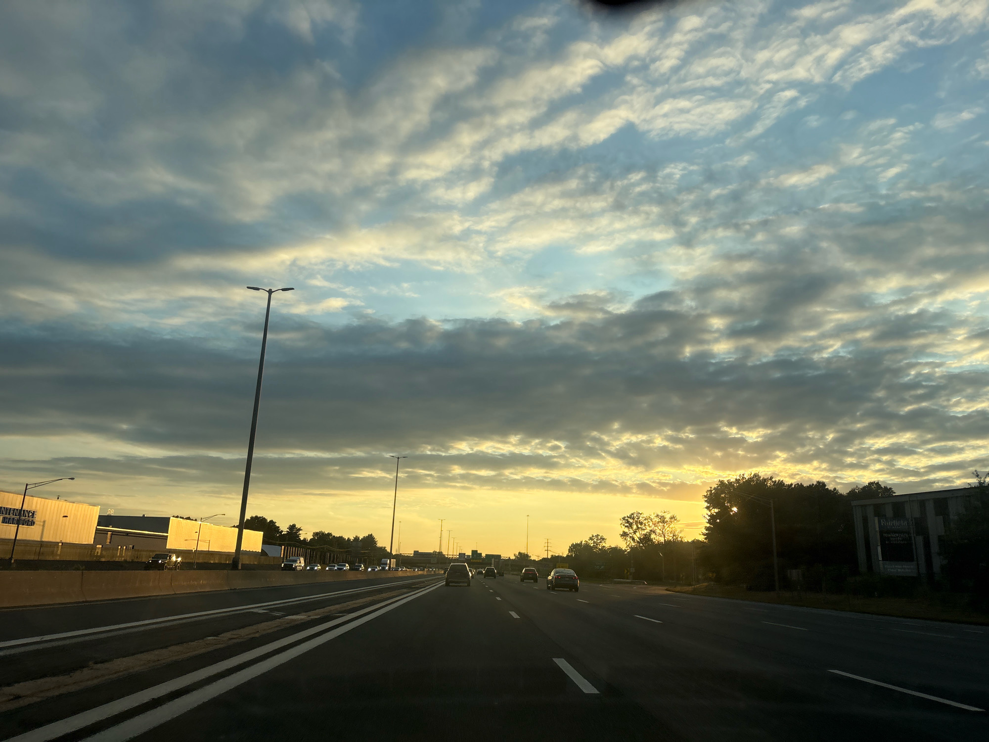 The image shows a view from a vehicle traveling on a highway in East Hartford, United States of America, at sunrise. The sky is filled with a dramatic display of clouds, with patches of sunlight breaking through, creating a gradient of light from yellow near the horizon to blue overhead. Tall streetlights line the highway. On the left side of the highway, there is a concrete barrier separating the lanes from a commercial building complex. The building appears to be a large, white industrial or warehouse structure with signage. On the right side of the highway, there are trees and another building visible. Several vehicles, including cars and a truck, are traveling on the highway in both directions. The road surface is asphalt with white lane markings. The overall scene conveys a sense of early morning travel and the beauty of a sunrise.