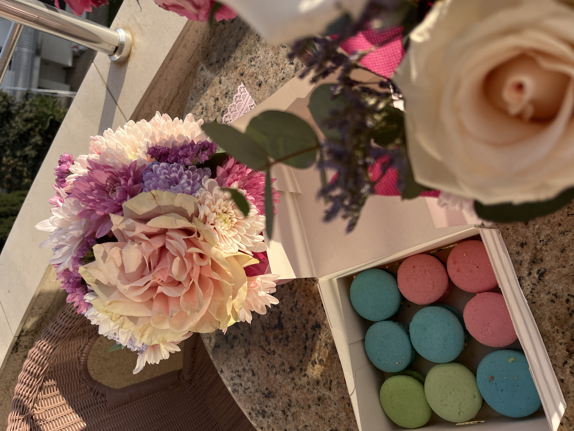 This media file captures a delightful outdoor still life scene, likely on a balcony or patio in Bečići, Montenegro. The primary subjects are two floral arrangements and a box of colorful macarons, resting on a speckled granite or marble-patterned surface.
In the foreground, a vibrant and substantial bouquet showcases a mix of flowers, predominantly in shades of pink, purple, white, and soft peach. Large, ruffled dahlia-like blooms in light pink are prominent, alongside dense clusters of chrysanthemums and smaller purple filler flowers. To the right of this bouquet, a white open box reveals nine macarons, uniformly colored in pastel light blue, pink, and light green, suggesting a thoughtful gift or treat. Further to the right and partially out of focus, a second, smaller floral arrangement features a soft, creamy white rose as its central piece, accompanied by delicate purple filler flowers and green foliage, including eucalyptus leaves.
The setting is bright and sunny, indicated by the strong, elongated shadows cast by the railing, flowers, and box onto the tabletop and a light-colored woven wicker chair or table base visible in the lower left. This directional light suggests either early morning or late afternoon. A polished metal railing or support structure is seen in the upper left, confirming an outdoor, elevated location. In the background, out of focus, are glimpses of green foliage and indistinct building structures, consistent with a resort or town environment in Bečići.
The overall impression is one of beauty, celebration, or a moment of enjoyment. No visible text is present on any objects in the frame.
