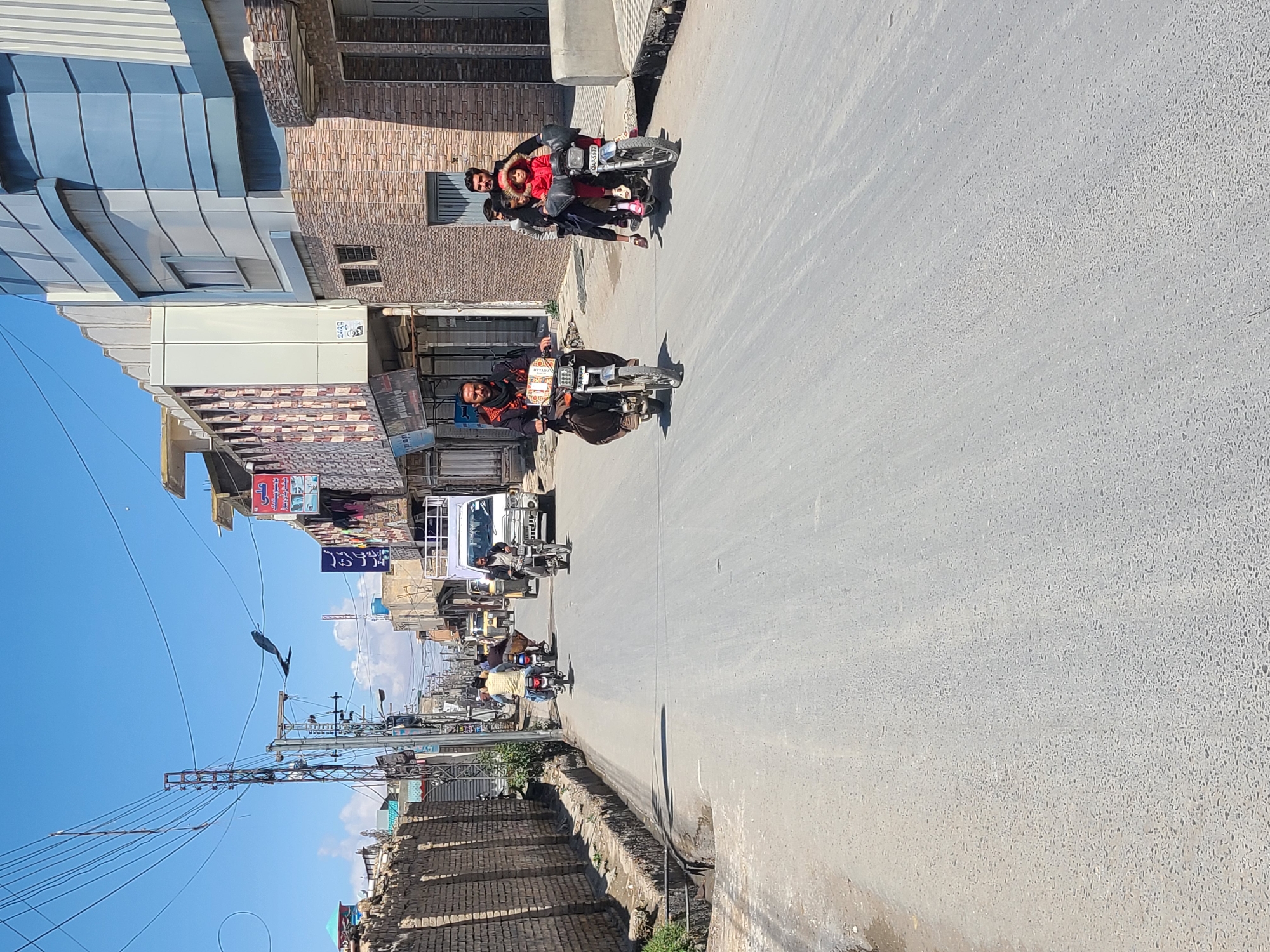The image depicts a street scene in Quetta City Tehsil, Pakistan. The view is looking down a road with buildings on either side. Motorbikes are prominent, with several people riding them. One motorbike has a rider and three passengers, one of whom is wearing a red coat. The sky is blue, and power lines run along the street. The buildings appear to be a mix of commercial and residential structures.