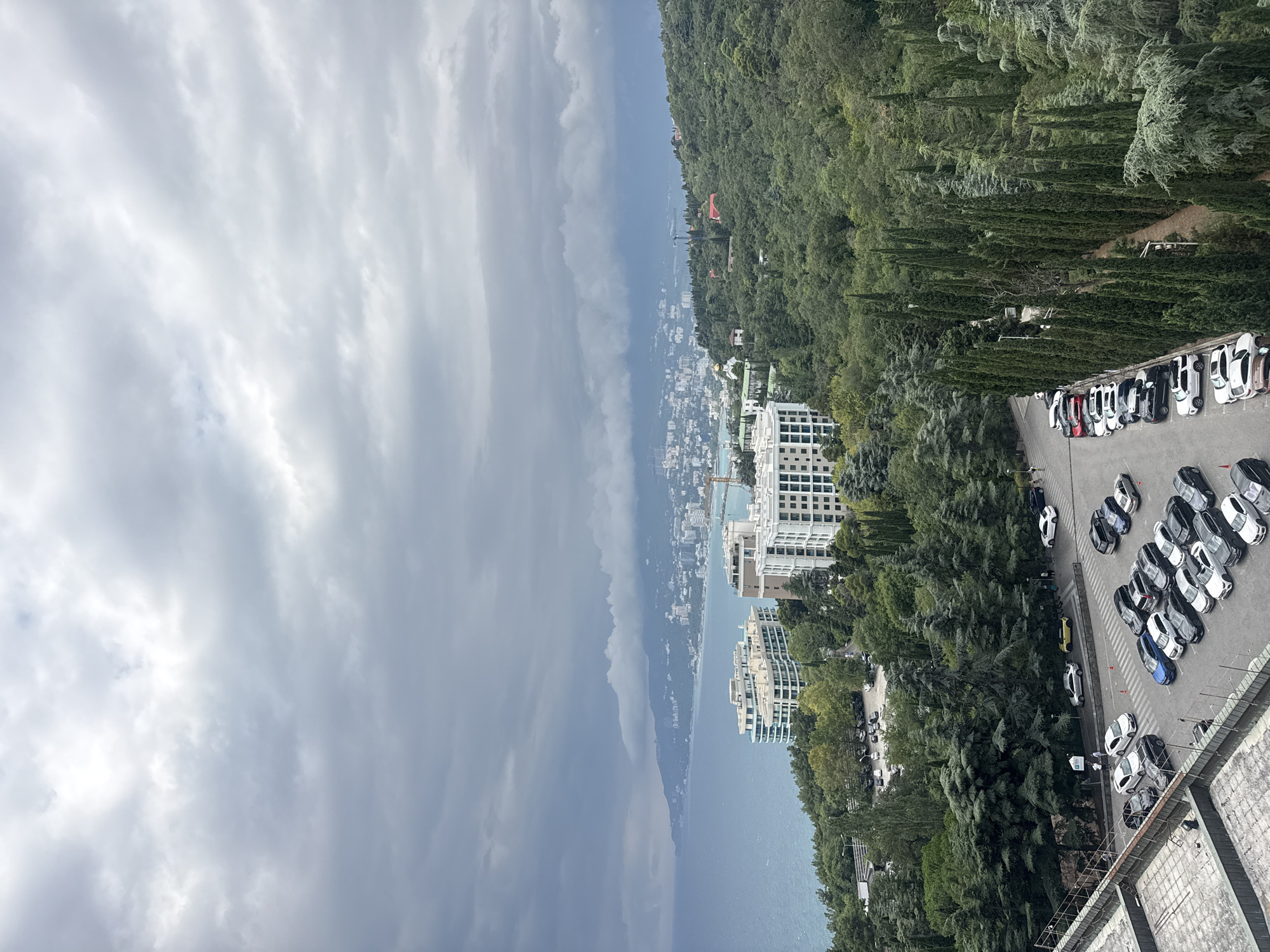 The image captures an elevated view of a coastal city, Yalta, Ukraine, under a cloudy sky. In the foreground, a large, light-colored building with a grid-like pattern of windows is visible, with its tiled roof and a metal railing. Adjacent to this building is a parking lot filled with numerous cars of various colors and models, including white, black, silver, blue, and beige sedans, SUVs, and hatchbacks. Some cars are parked in marked bays, while others are more loosely arranged.
Beyond the parking lot, dense green trees and cypress trees create a lush forest. A road runs through the trees, and a few cars are parked along its edge. Further in the background, more modern buildings, including white, multi-story structures with a distinctive curved design, are situated closer to the coast. These buildings appear to be hotels or apartment complexes.
The coastline stretches into the distance, meeting the calm, blue sea under a heavy, overcast sky with thick clouds. The distant cityscape of Yalta is visible along the coast, with a mix of buildings and infrastructure extending towards the mountains. The overall scene suggests a somewhat overcast day in a scenic coastal city with significant urban development amidst natural greenery. No people are prominently featured or engaged in any specific actions in the main part of the image.