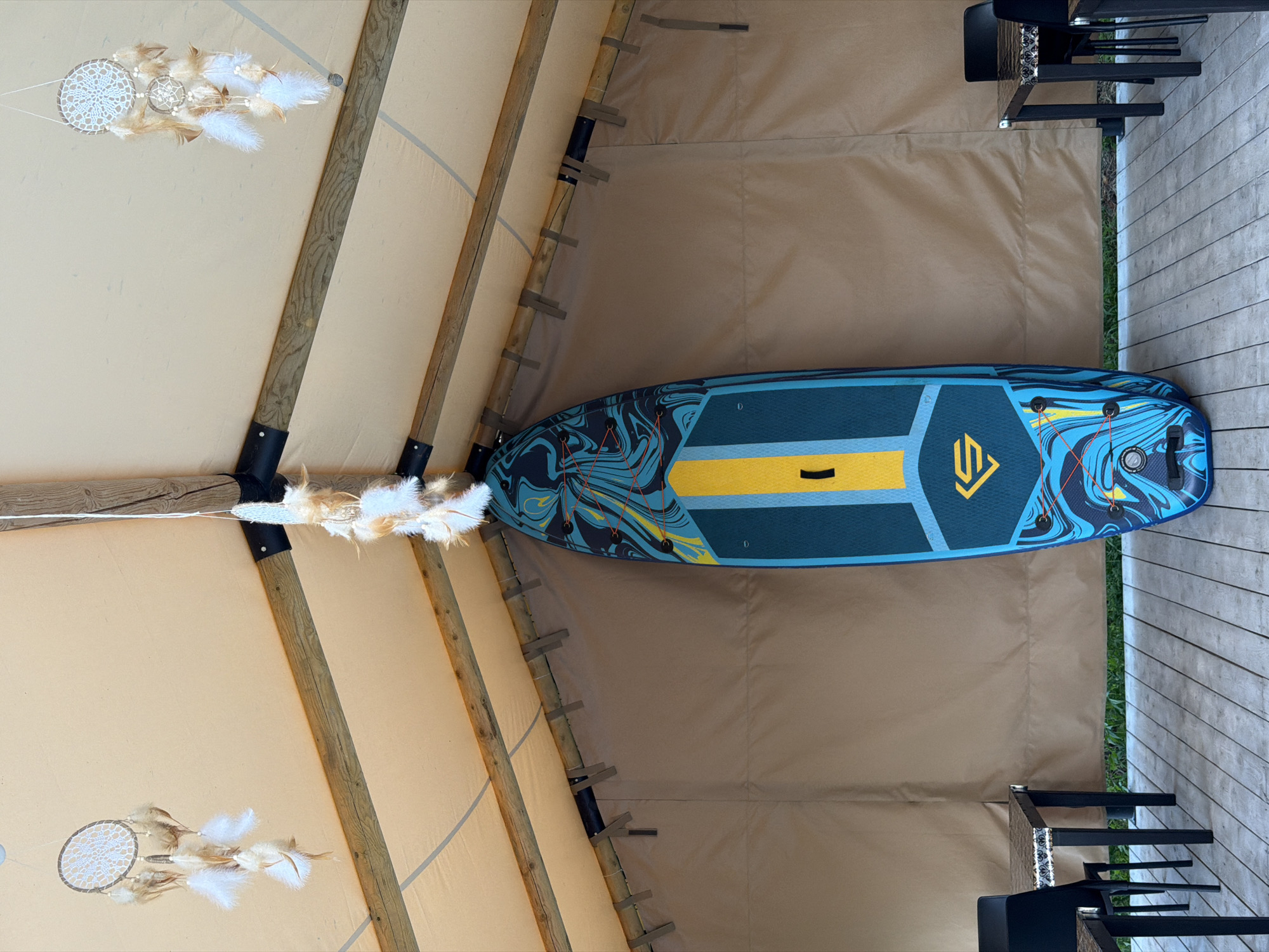 This image depicts the interior of a tent-like structure with a beige canvas roof supported by wooden beams. A stand-up paddleboard with a blue, yellow, and black abstract design is hanging vertically from the ceiling in the center of the frame. Two dreamcatchers with white feathers hang from the ceiling, one on the left and one on the right. The walls of the structure are also made of beige canvas. In the foreground, a wooden deck is visible, and on either side of the image, black chairs and tables are placed. The scene appears to be a covered outdoor seating area, possibly part of a glamping site or a recreational facility. The specific location in Esso, Russia, is not visually indicated within the image itself.