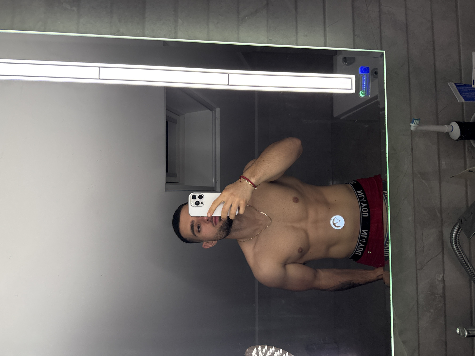 A shirtless man stands in front of a mirror, taking a selfie with a white iPhone. He is wearing red underwear with a black waistband that has white lettering. He has a gold chain around his neck and a red bracelet on his left wrist. The mirror has a light at the top and is mounted on a grey tiled wall. A white cabinet is visible in the mirror's reflection. An electric toothbrush holder and a shower hose are attached to the wall to the right of the mirror. The man appears to be in a bathroom. The location in the city of Gabrovo, Bulgaria cannot be determined.