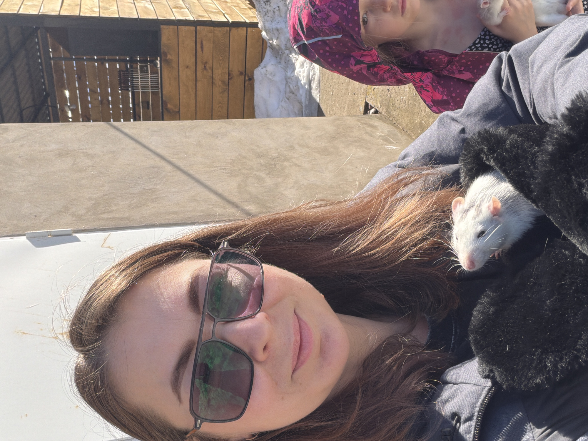 An outdoor scene depicts two females with two white domestic rats under bright sunlight. An adult female, positioned in the foreground and left, has long brown hair, wears dark-framed sunglasses, and a dark jacket. She holds one white domestic rat in her black-mitten-covered hands and smiles slightly towards the camera. Partially visible on the right, behind the adult, is a younger female wearing a pink patterned hood and a dark top, holding a second white domestic rat in her right arm. The background includes a light-colored wall or door, a wooden slatted structure with a dark roof, and a pile of snow on the ground. The ground is paved or concrete. The location is an unidentifiable outdoor area in Unknown, Unknown.