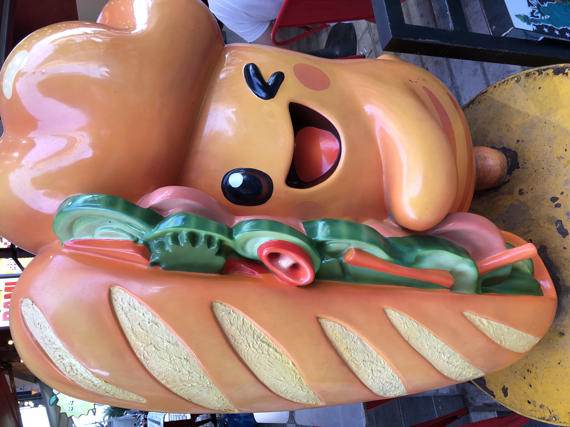 An outdoor sculpture of a cartoon hot dog is displayed at a street food stall. The sculpture is orange and yellow, with a pink bun and green pickles. A person wearing a white t-shirt is partially visible in the background, seated at a red chair. The sculpture is positioned on a yellow, weathered metal table. In the distance, signs with Vietnamese text are visible, indicating the location as Đà Nẵng, Vietnam. The ground is paved with gray bricks.