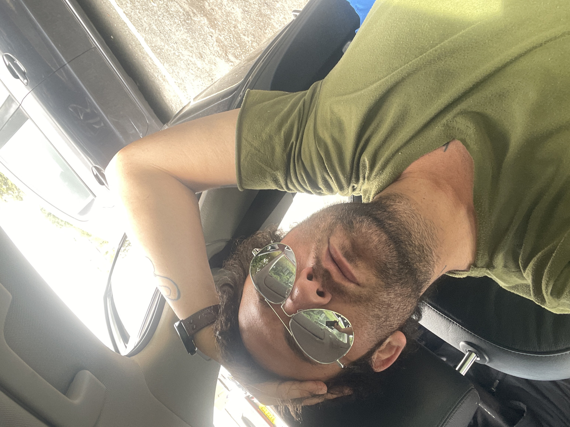 A man, appearing to be in his late 20s or early 30s, is pictured from a first-person perspective inside a vehicle. He is wearing a green t-shirt and aviator sunglasses. His right arm is raised, with his hand resting on his head, and his left arm is bent, with his hand on his lap. The man has dark, curly hair and a beard. The interior of the car is visible, including a black leather seat and a sunroof that is partially open, allowing sunlight to stream in. Outside the car, a gray vehicle is parked. The overall lighting suggests it is daytime. The scene does not provide any specific details to determine the location within the city of Armagh, United Kingdom.