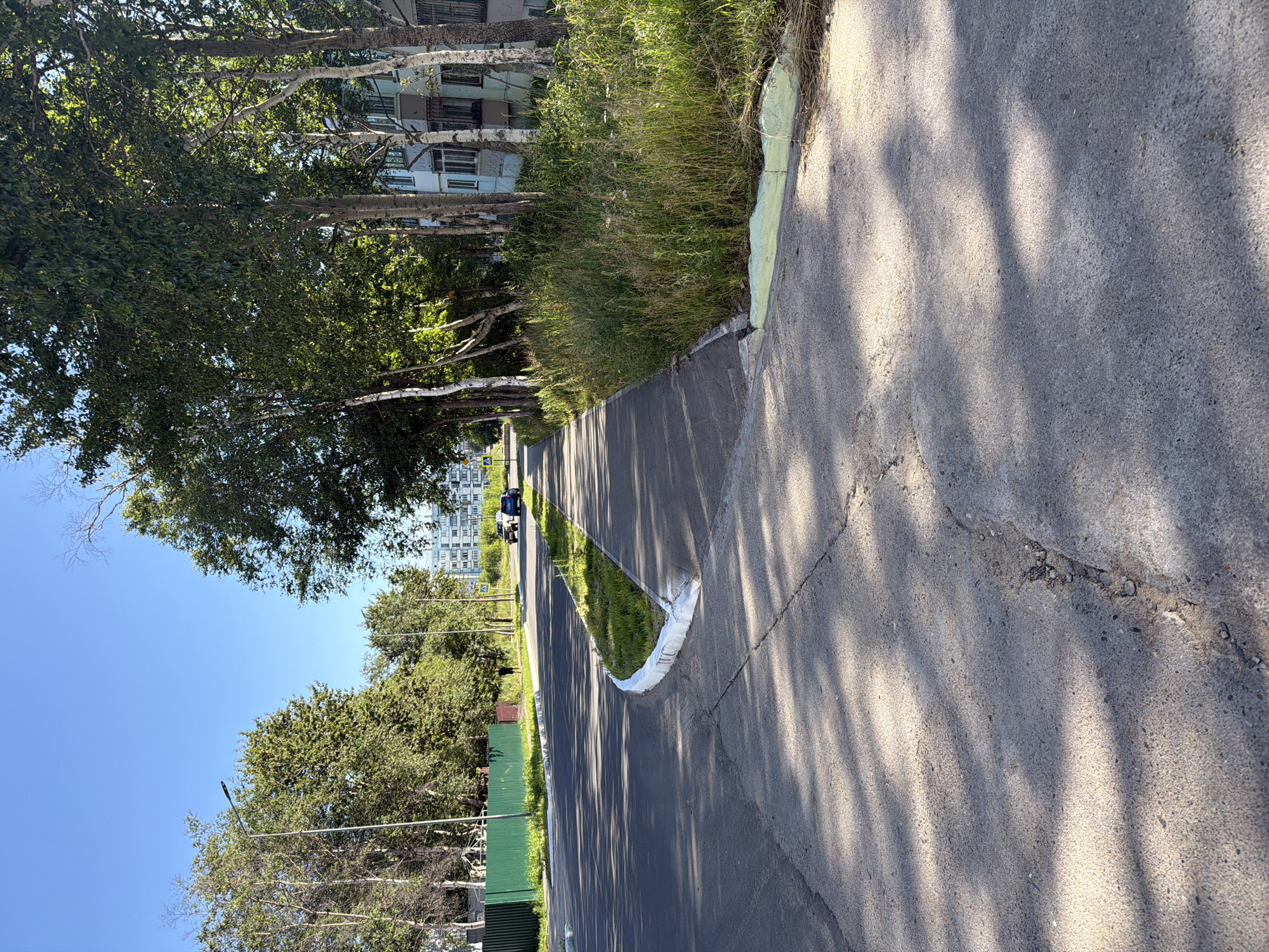 The image depicts a street scene in the city of Okha, Russia, on a clear, sunny day. A paved road runs diagonally through the frame, with a concrete sidewalk bordering it on the right. The sidewalk is shaded by a row of trees, their leaves a vibrant green. Beyond the trees, apartment buildings with light-colored facades are visible. On the left side of the road, a green fence encloses an area with some structures, possibly a playground. Further down the road, two cars are driving away from the camera, and a lone pedestrian is walking on the sidewalk. The ground in the foreground shows signs of wear and tear, with cracks and shadows from the trees cast across the asphalt. The overall impression is of a typical urban street scene in a northern climate during the summer.