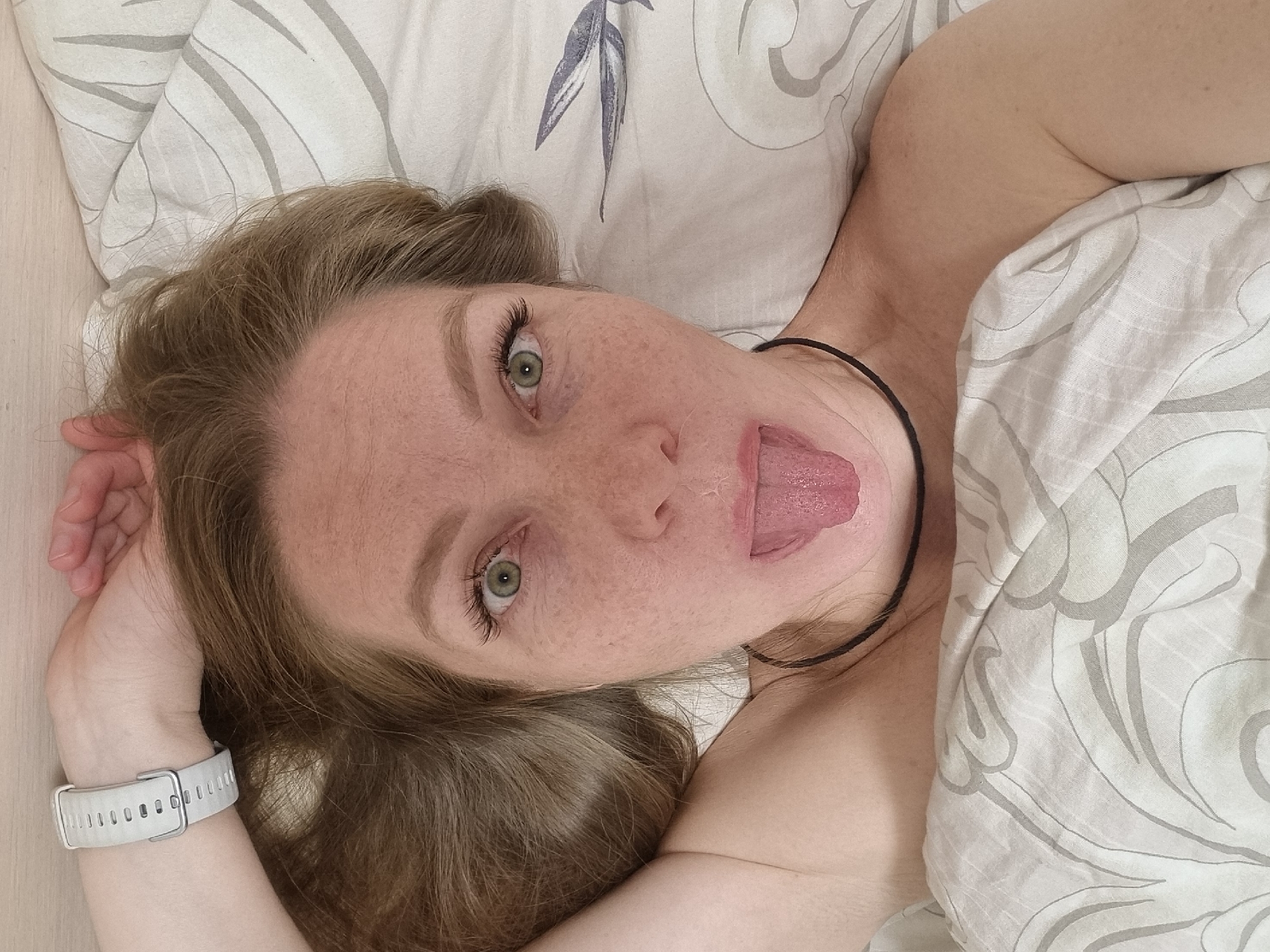 A woman with freckles and light brown hair is lying on patterned bedding, sticking out her tongue. She is wearing a white smartwatch. The location is unknown.