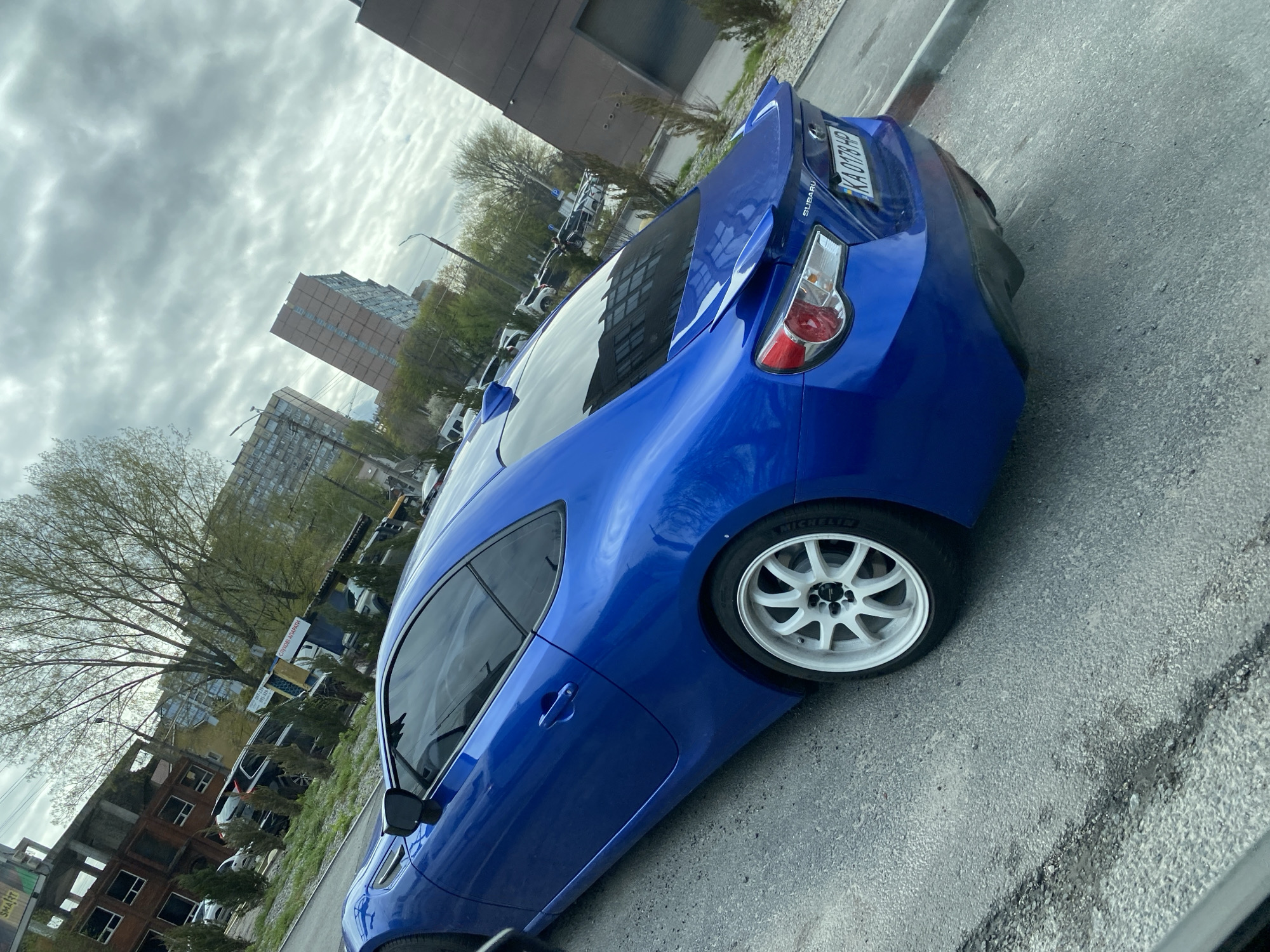 The image shows a blue Subaru sports car parked on an asphalt surface. The car has white aftermarket wheels and a rear spoiler. In the background, there are buildings, trees, and other parked vehicles. The sky is overcast. The license plate of the car appears to be KA 0178 HP. The image appears to be taken in an urban environment in the city of Unknown, Unknown.