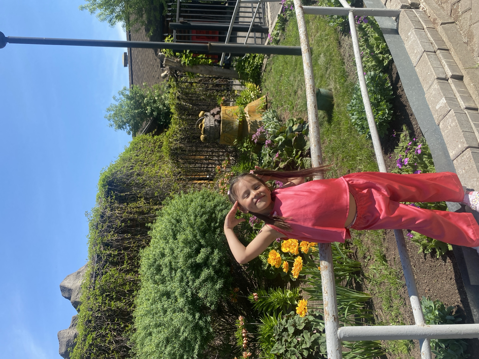 A young girl in pink pajamas stands by a metal railing in an outdoor garden setting on a sunny day. The garden includes flowering plants, hedges, and a wooden sculpture. In the background, there is a house with a dark roof. The location is presumed to be Izhevsk, Russia.