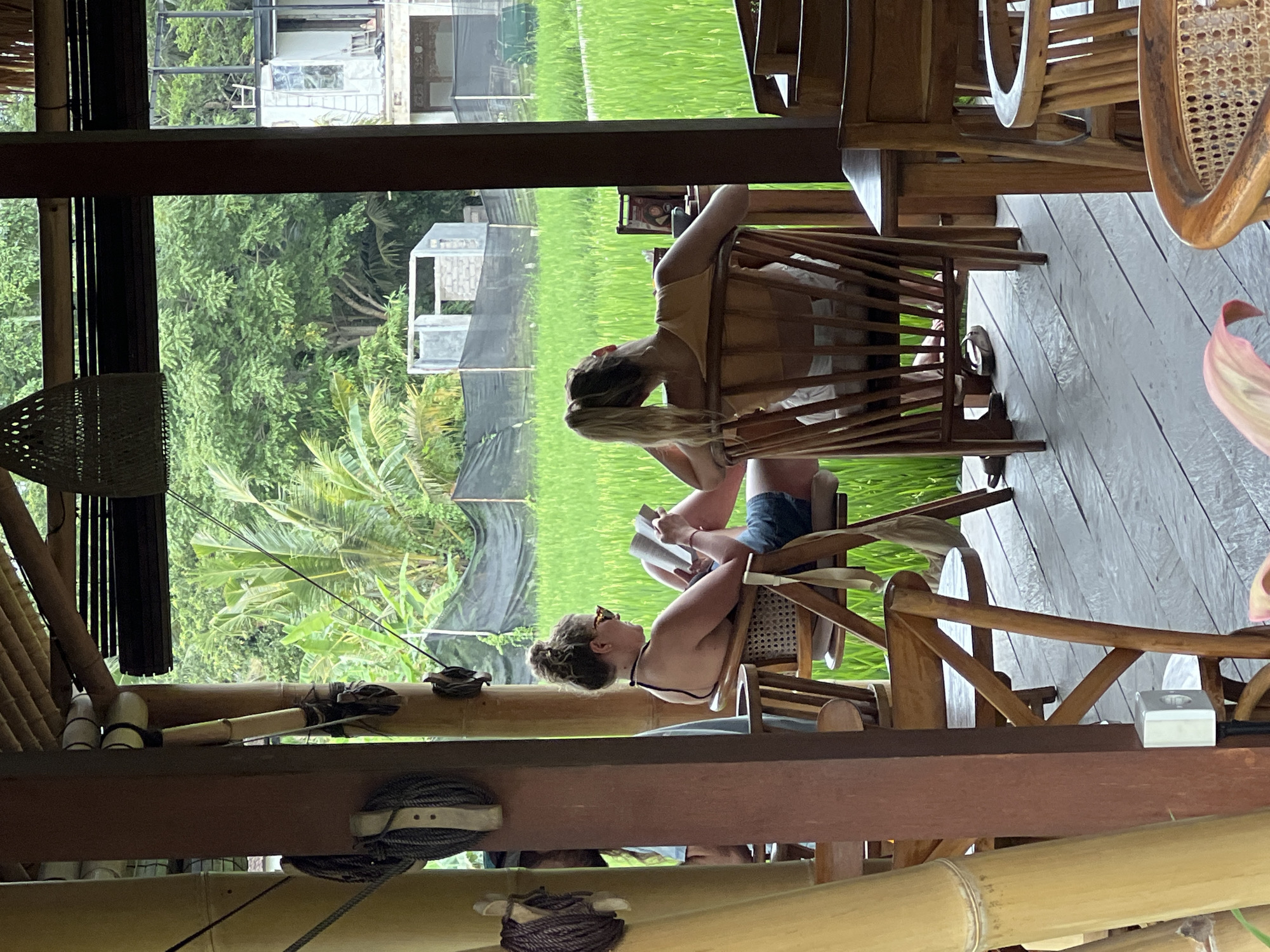 Two women are seated on wooden chairs, potentially made of rattan, within a structure predominantly composed of bamboo. They are positioned next to a rice field landscape in Ubud District, Indonesia. One woman is reading a book, while the other is reclining back in her chair. The structure they are within appears to be a cafe or restaurant, based on the seating arrangement and overall design.
